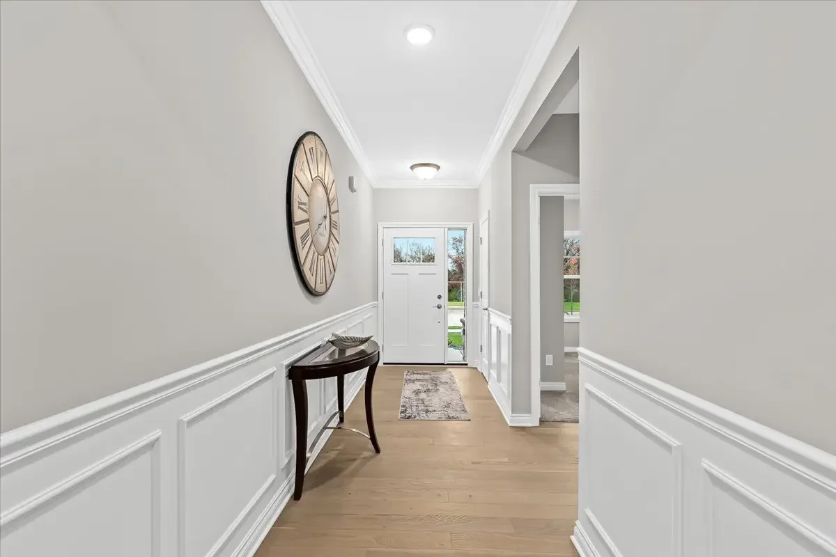 Empty entryway with white wainscoting, light gray walls, hardwood floor, a small dark wood table with decorative bowl, large round wall clock, front door with window, and a view of outdoors.