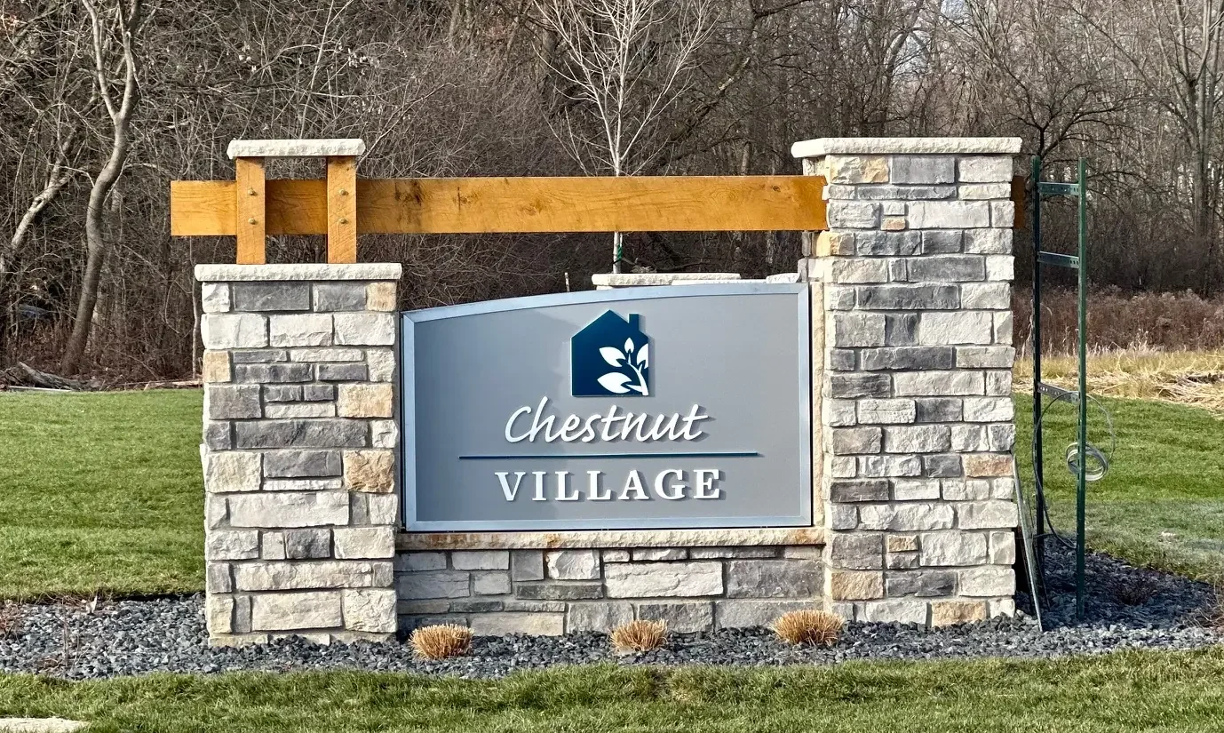 Entrance sign for Chestnut Village, built into a stone brick wall with a metal sign featuring a house logo and text, surrounded by well-maintained grass and leafless trees in the background.
