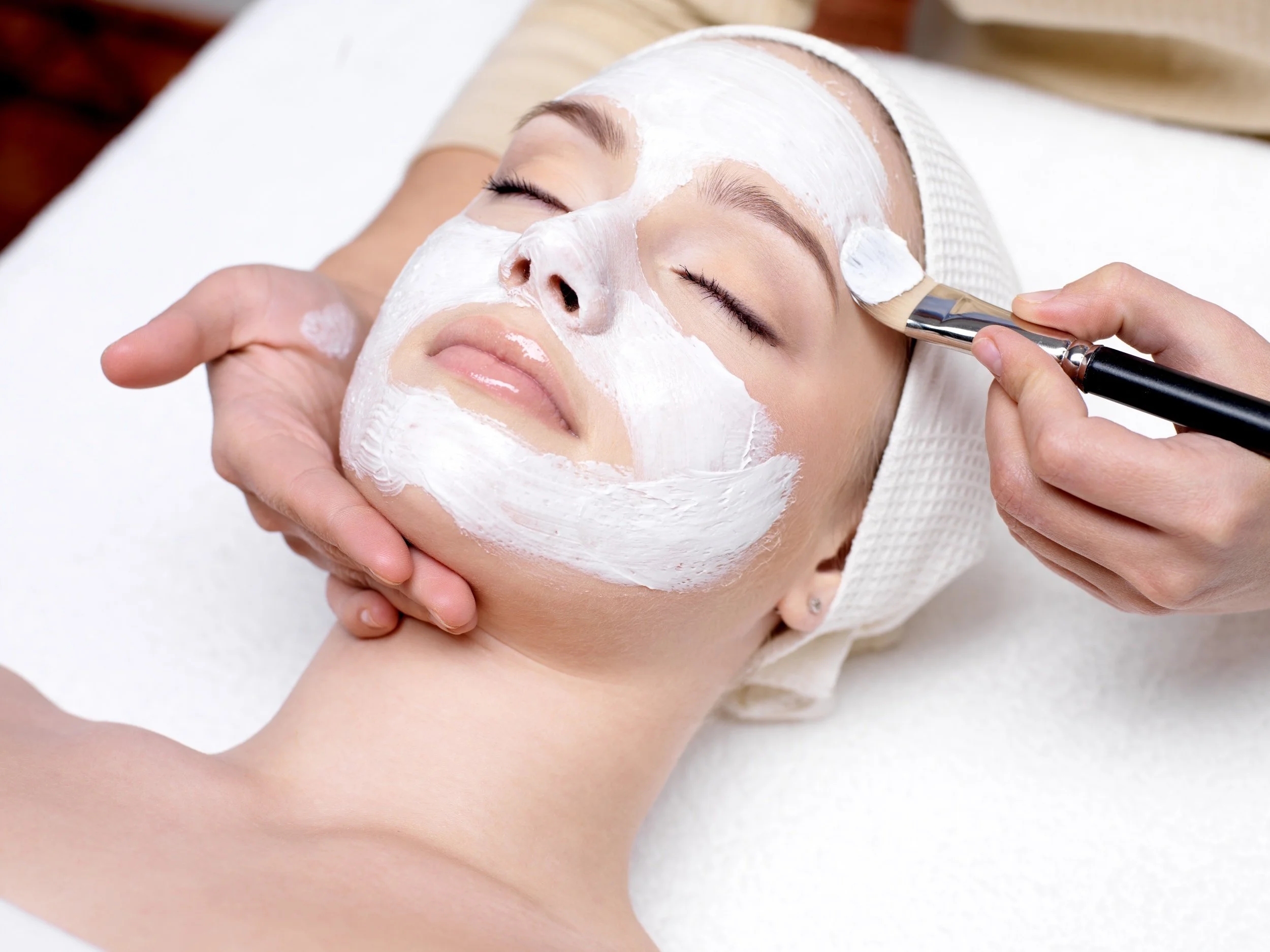 Une femme reçoit un soin du visage avec une crème blanche appliquée sur son visage, tout en étant relaxée, dans un salon de beauté.