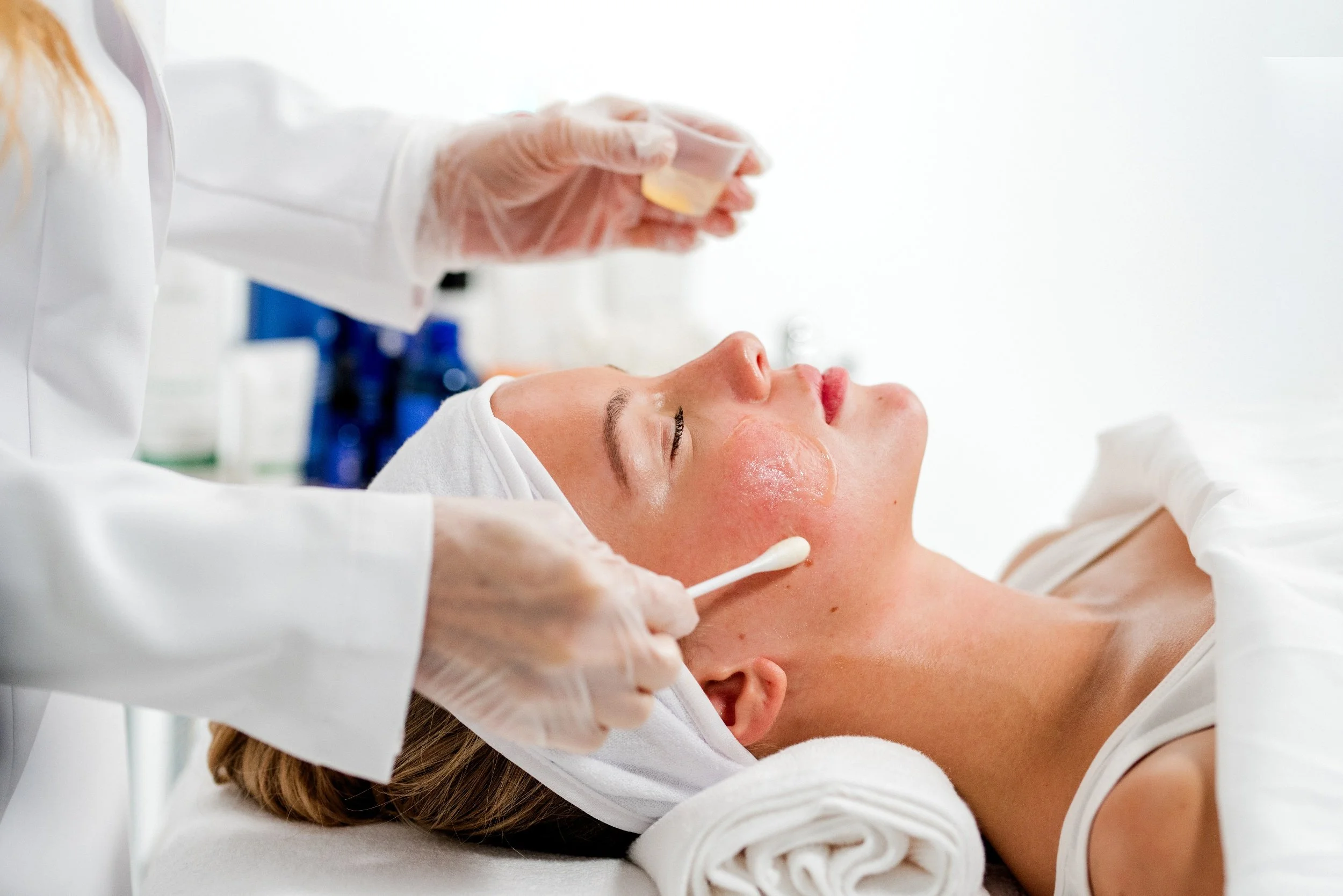 Une femme reçoit un soin du visage dans un spa ou un centre de beauté, allongée avec une serviette sur la tête, pendant qu'une esthéticienne applique un produit sur son visage.