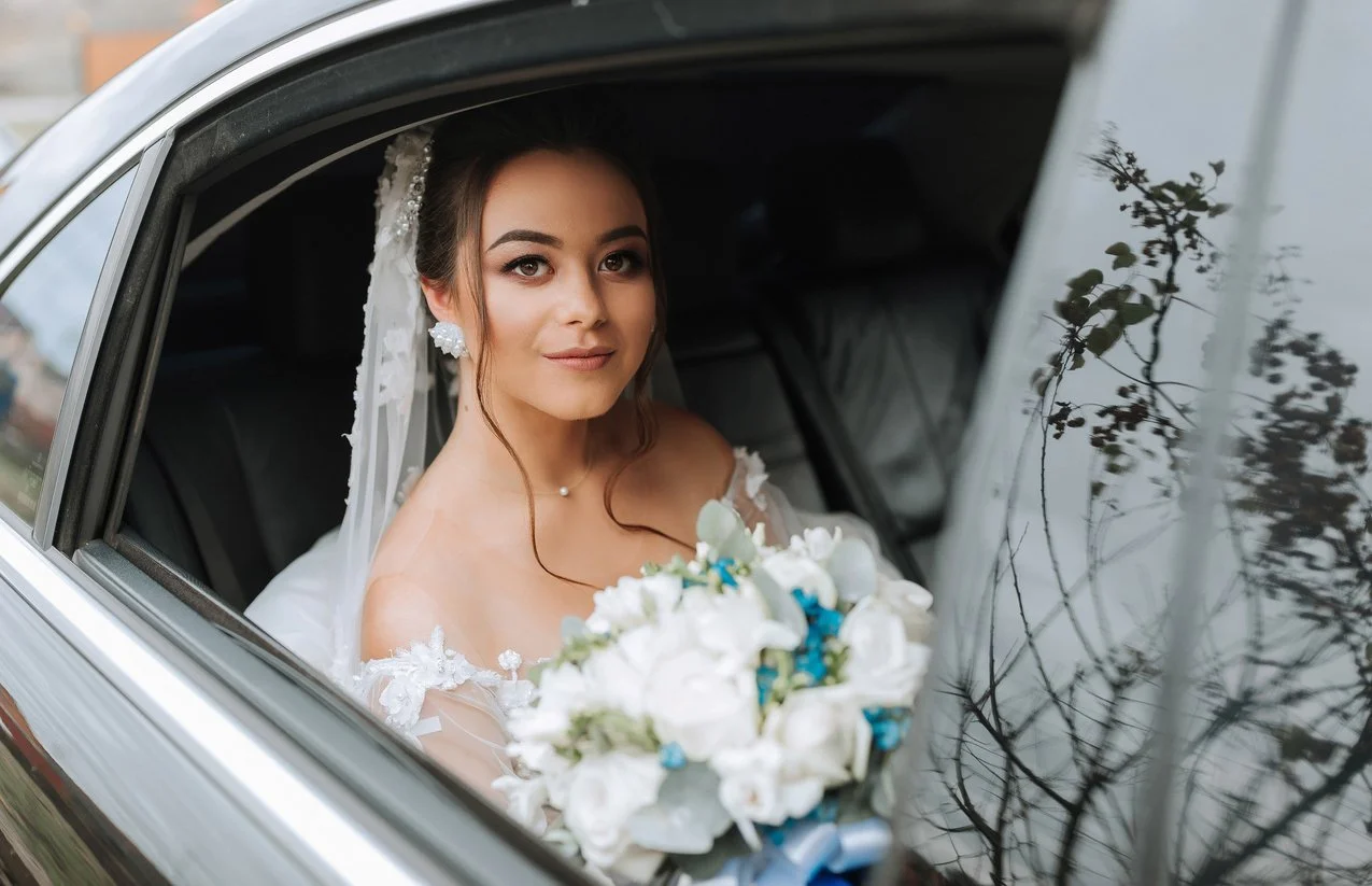 A bride sitting inside a car, looking out the window, holding a bouquet of white and blue flowers, wearing a wedding dress and veil with floral details.