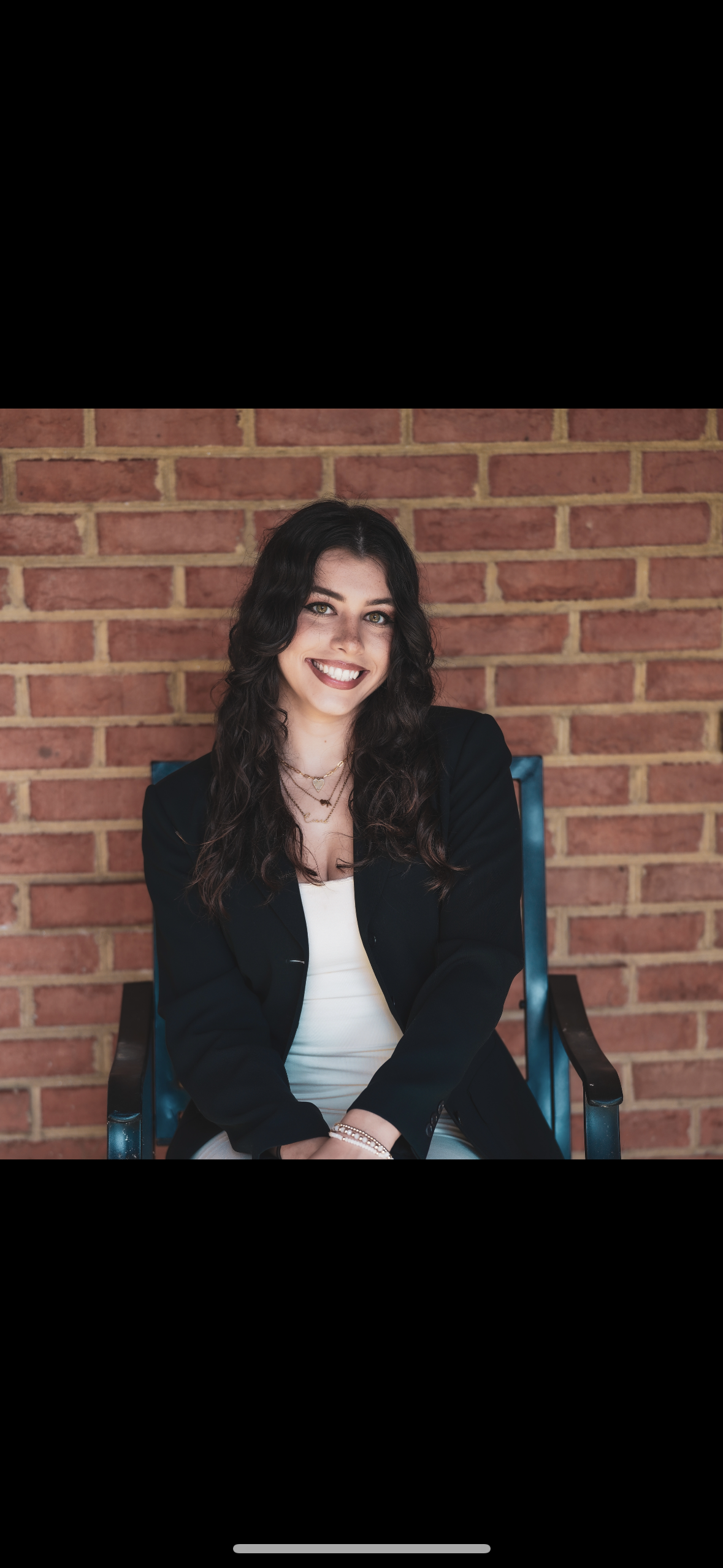 Young woman with curly dark hair, wearing a black blazer, sitting on a chair in front of a brick wall, smiling, with layered necklaces and a white top.