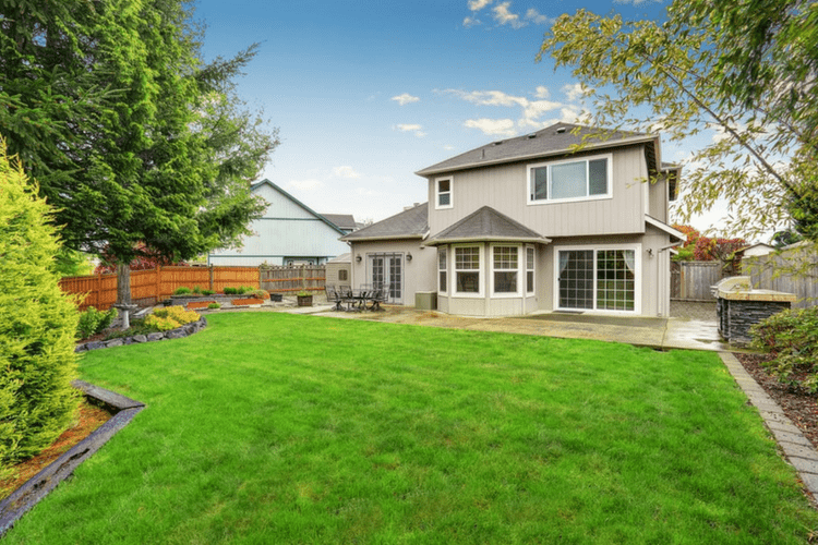 A backyard with a lush green lawn, trees, a patio area with outdoor furniture, and a two-story house with large windows.