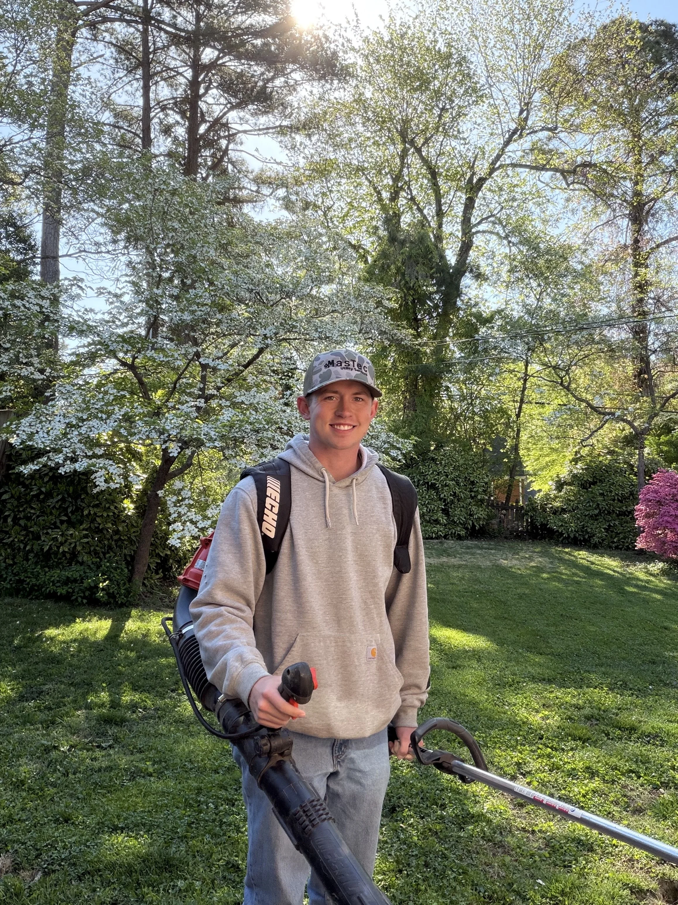 A young man with a gray hoodie, a backpack, and a baseball cap smiling in a sunlit garden holding a leaf blower.