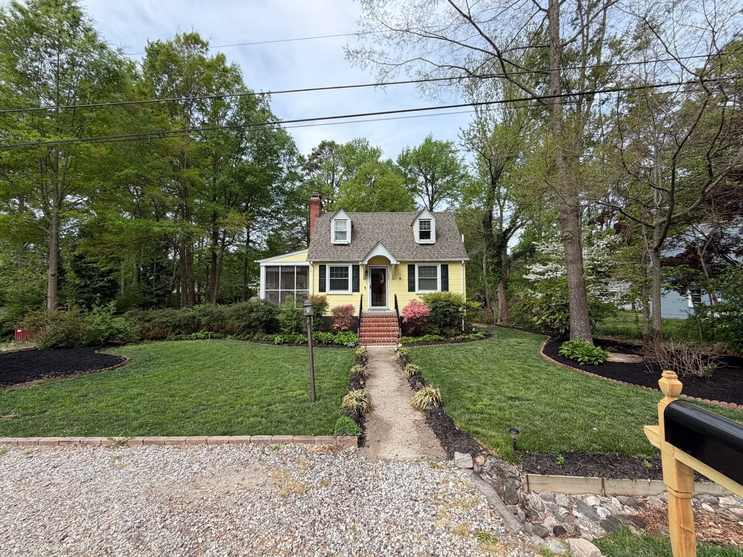 A yellow house with a porch and two dormer windows, surrounded by a landscaped yard with bushes, trees, and a gravel driveway.