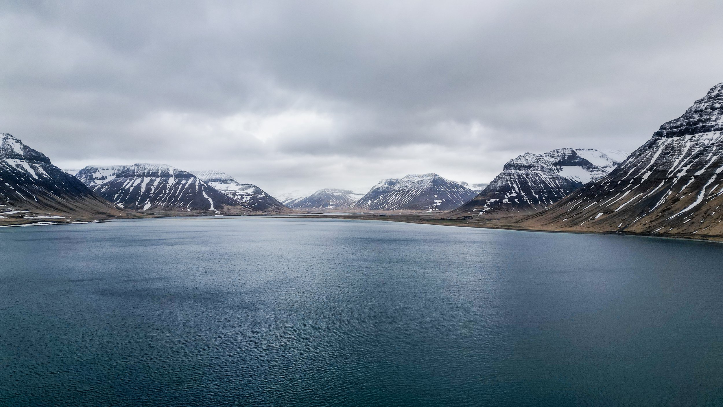 A landscape featuring a large body of water surrounded by snow-capped mountains under a cloudy sky.