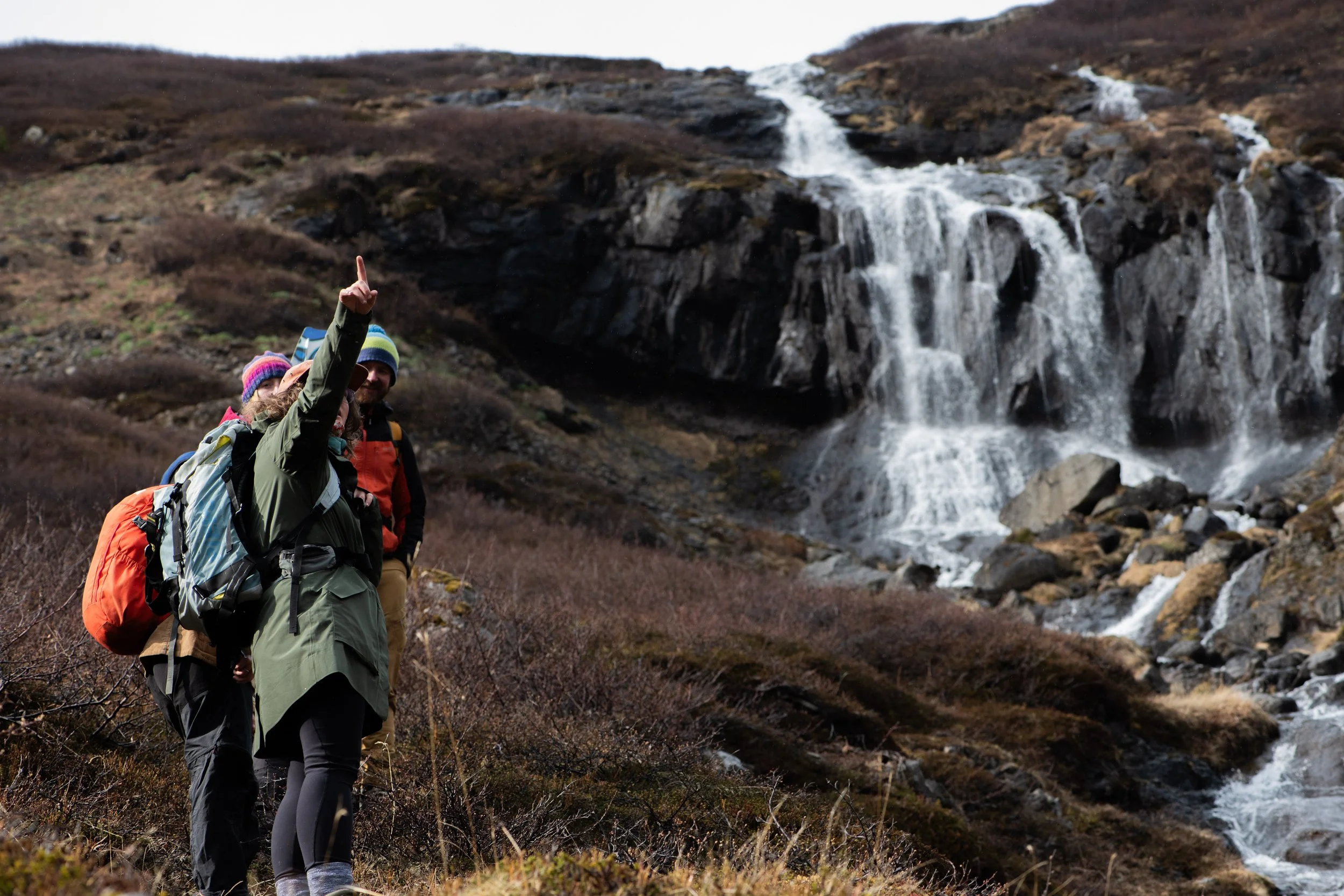 Hikers pointing towards a mountain waterfall with rugged terrain and brown bushes.