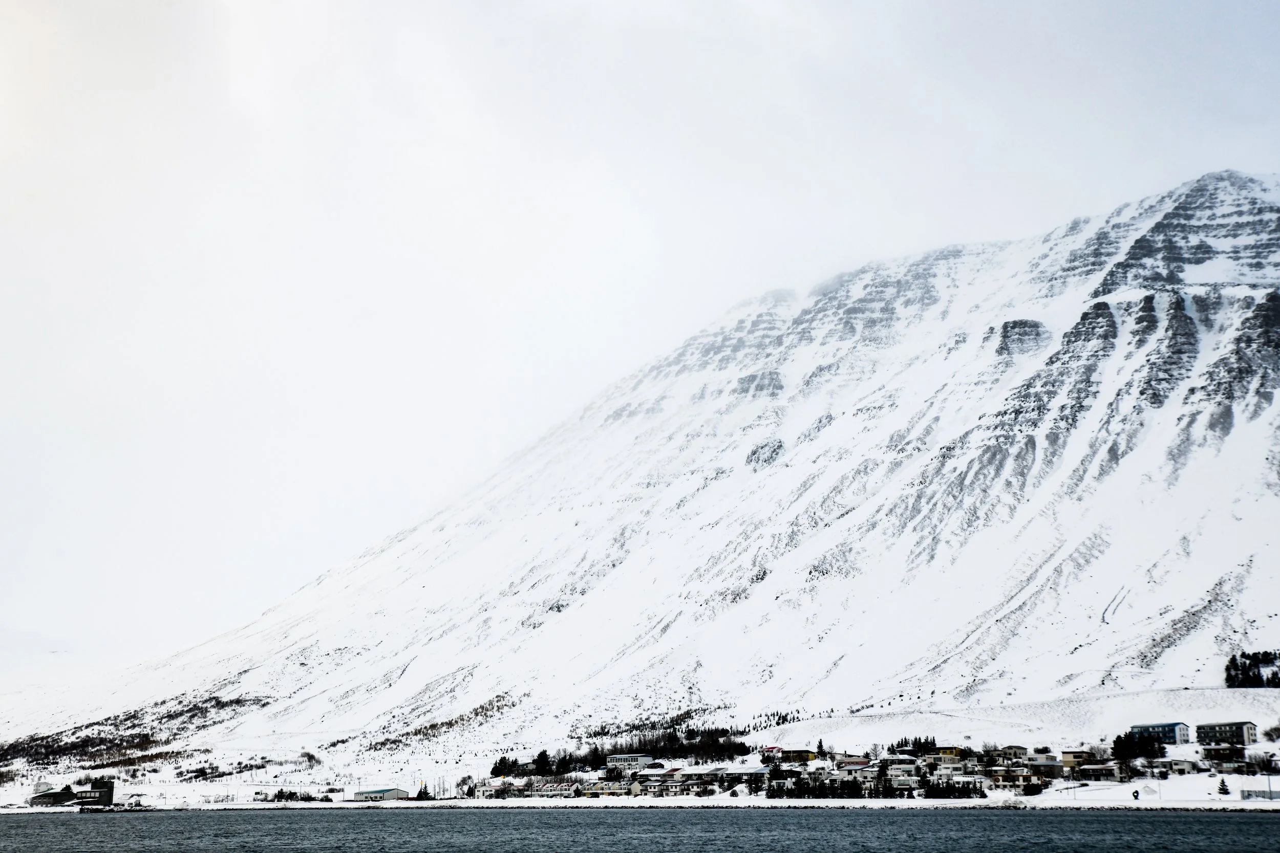 Snow-covered mountain with a small residential area at the base and a body of water in the foreground.