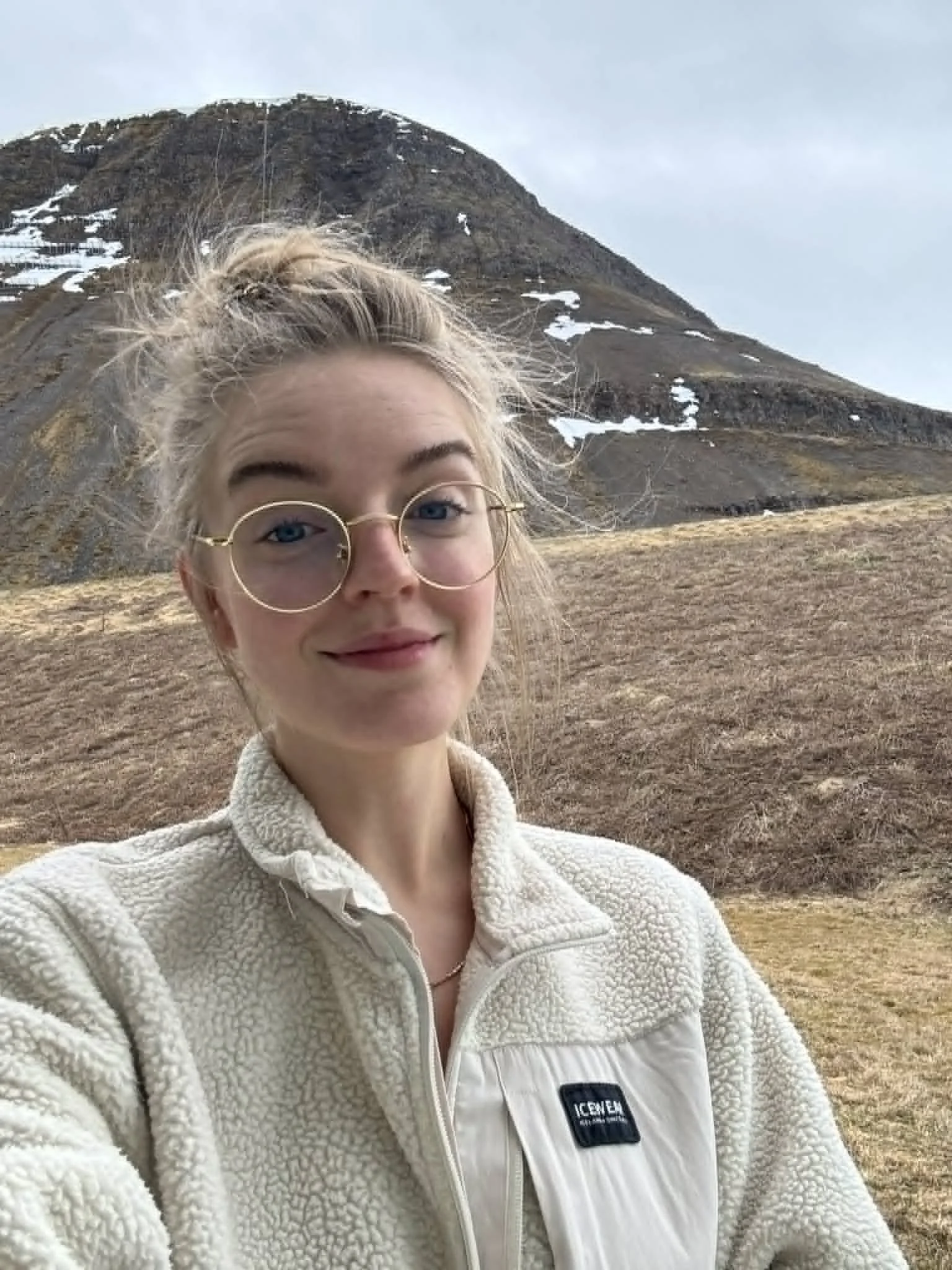 Close-up selfie of a young woman with glasses and blonde hair in a bun, standing outdoors in front of a mountain with patches of snow, grassy terrain, and a cloudy sky.