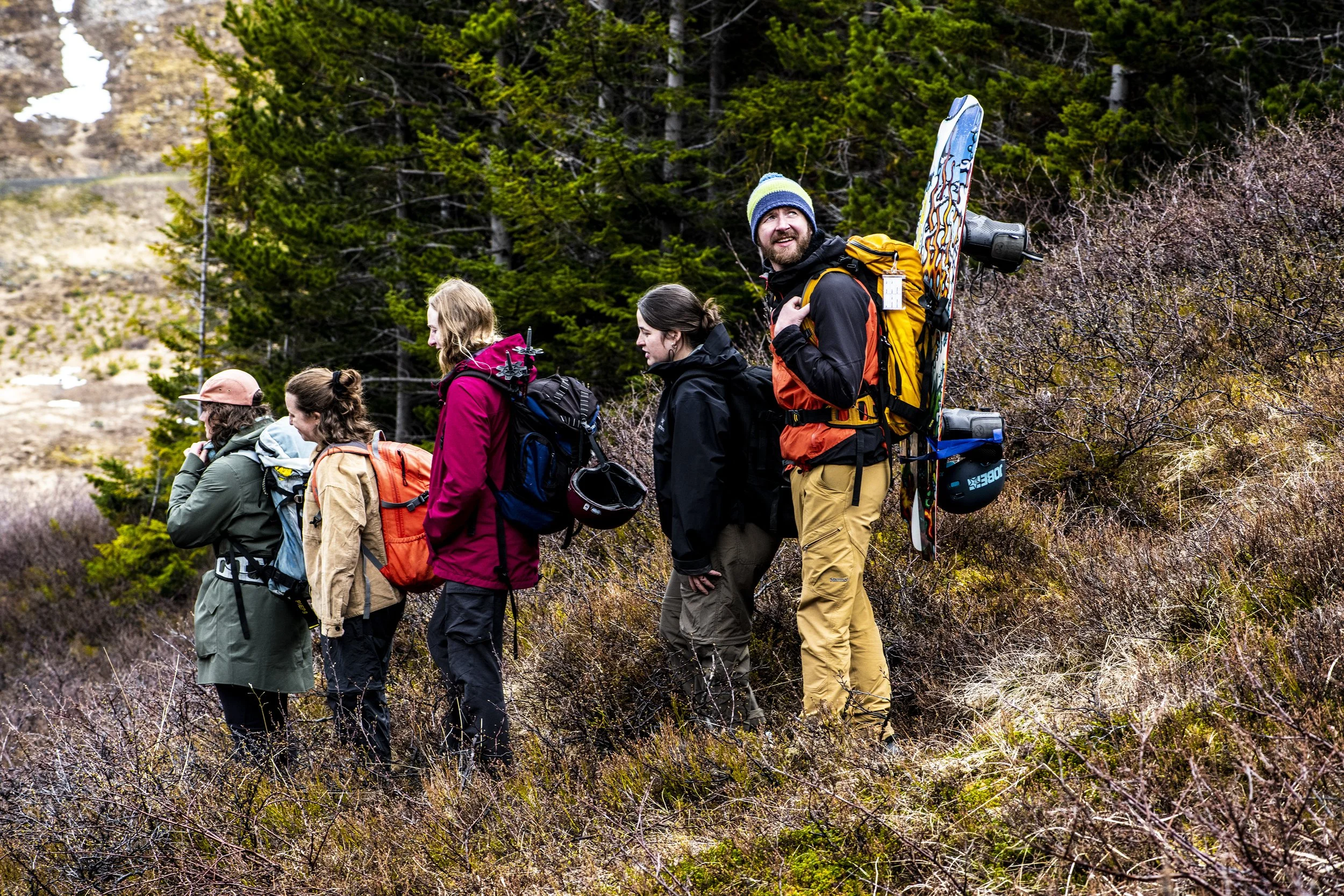 A group of hikers walking in a single file through a forested mountainous area, wearing outdoor gear and backpacks.