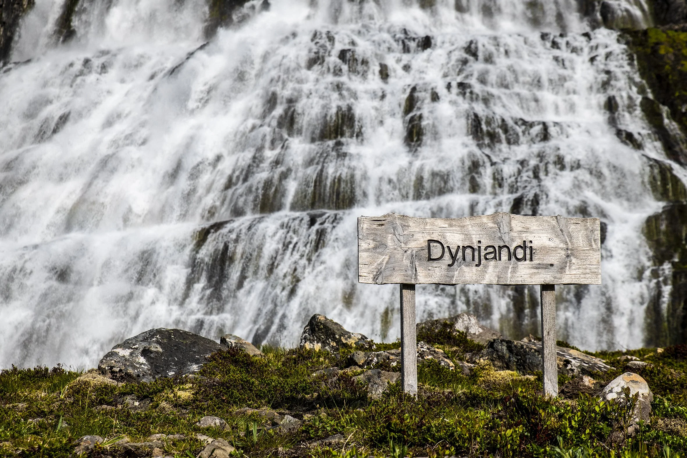 A wooden sign reading 'Dynjandi' in front of a large cascading waterfall with water flowing over rocks and moss on the ground.