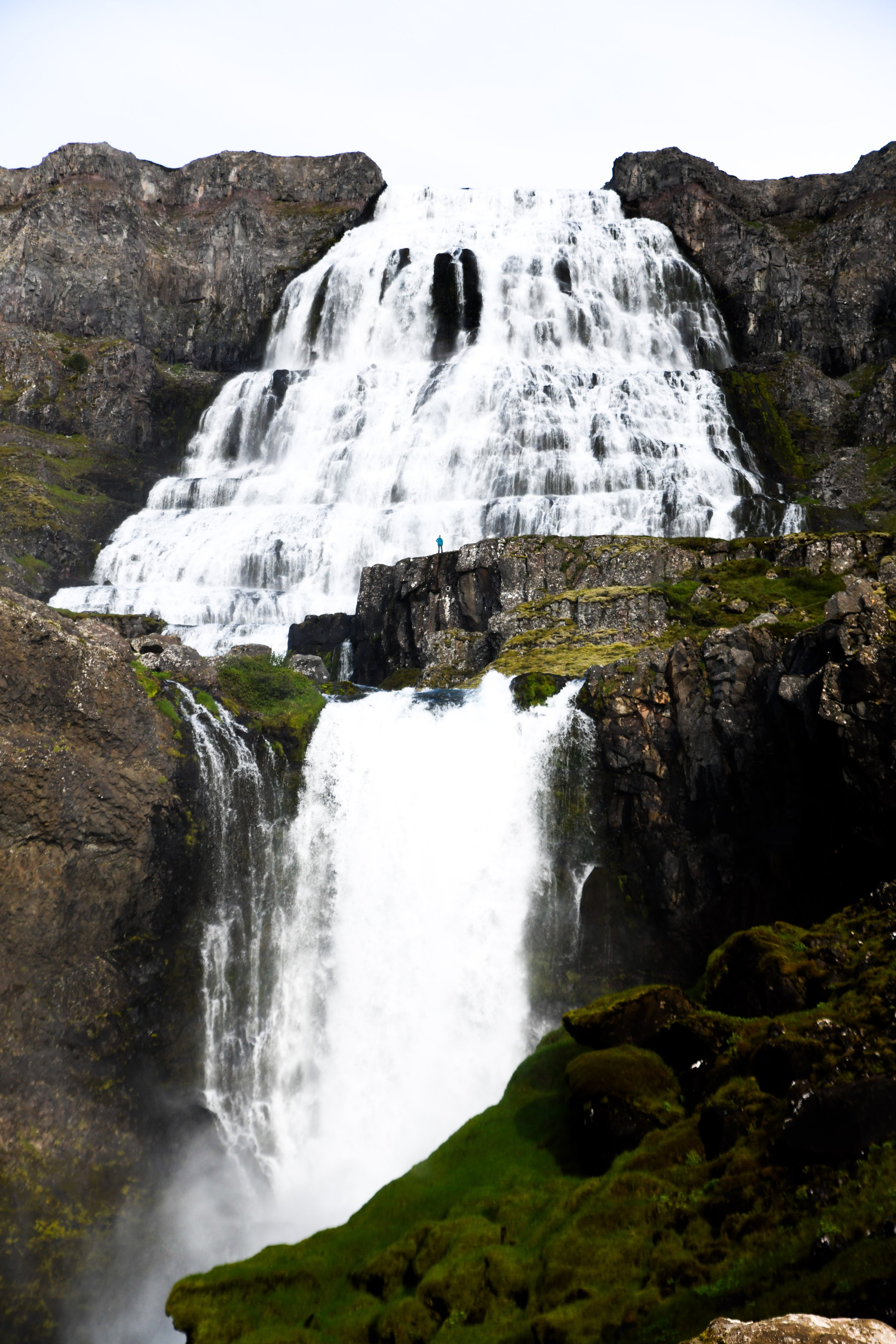 A tall cascading waterfall flowing over dark, rugged rocks with patches of green moss, set against a cloudy sky.