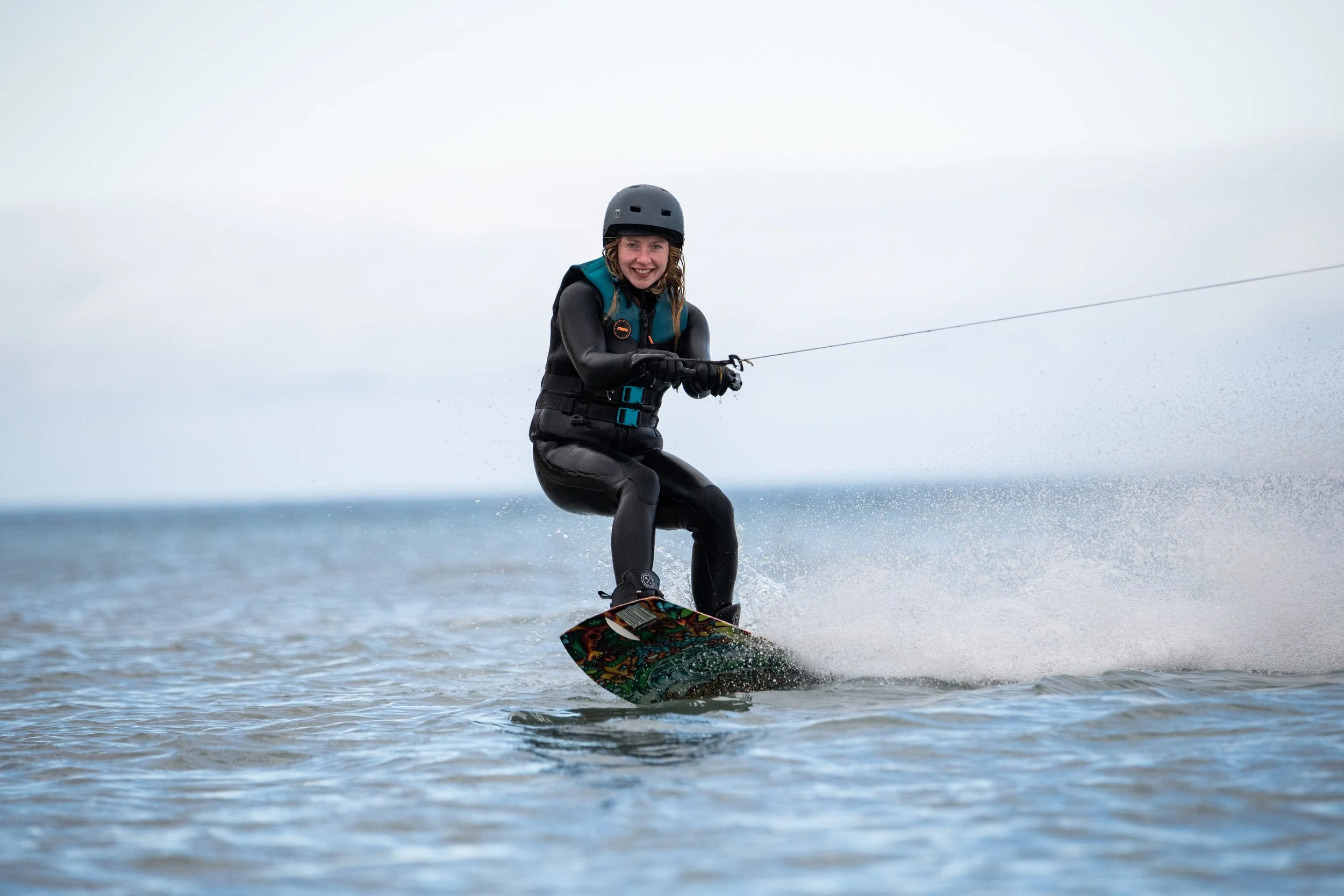 A woman wearing a helmet and wetsuit wakeboarding on a body of water, holding onto a rope with a smile on her face during overcast weather.