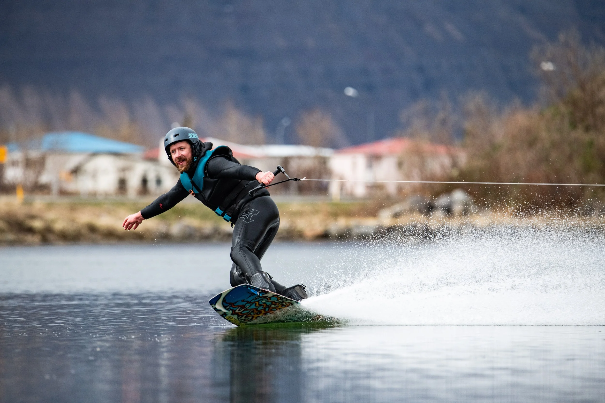 A man wakeboarding on a lake, wearing a helmet and life jacket, with houses and trees in the background and mountains in the distance.