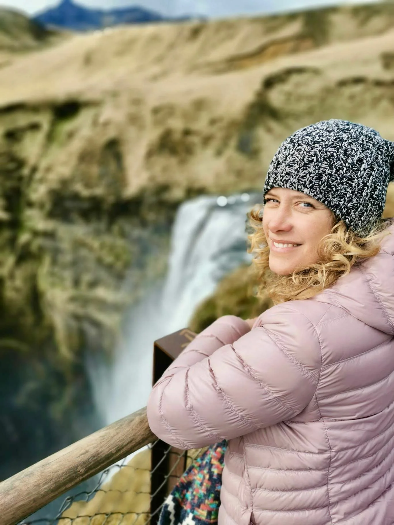 Woman with blonde curly hair, wearing a black and white knit beanie and a pink puffer jacket, smiling and looking at the camera while sitting by a wooden railing on a bridge overlooking a waterfall in a canyon.