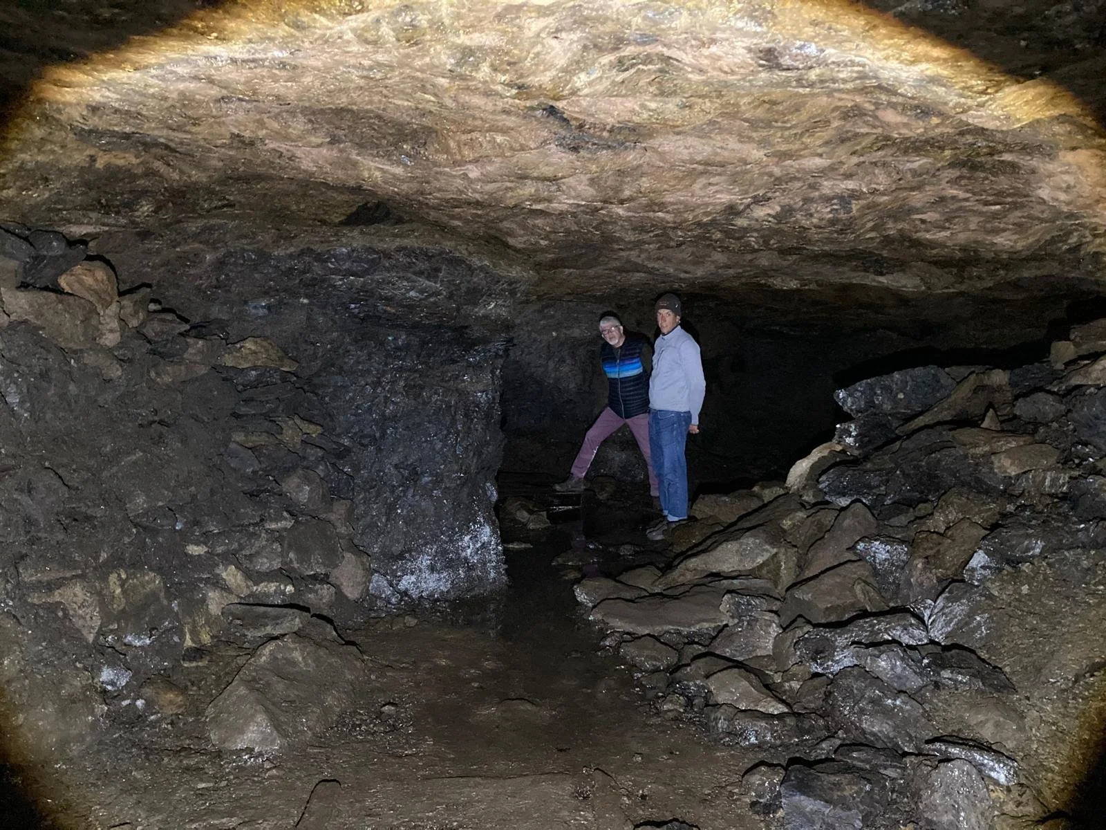 Two men standing inside a dark cave with rocky walls and ceiling, and uneven ground.