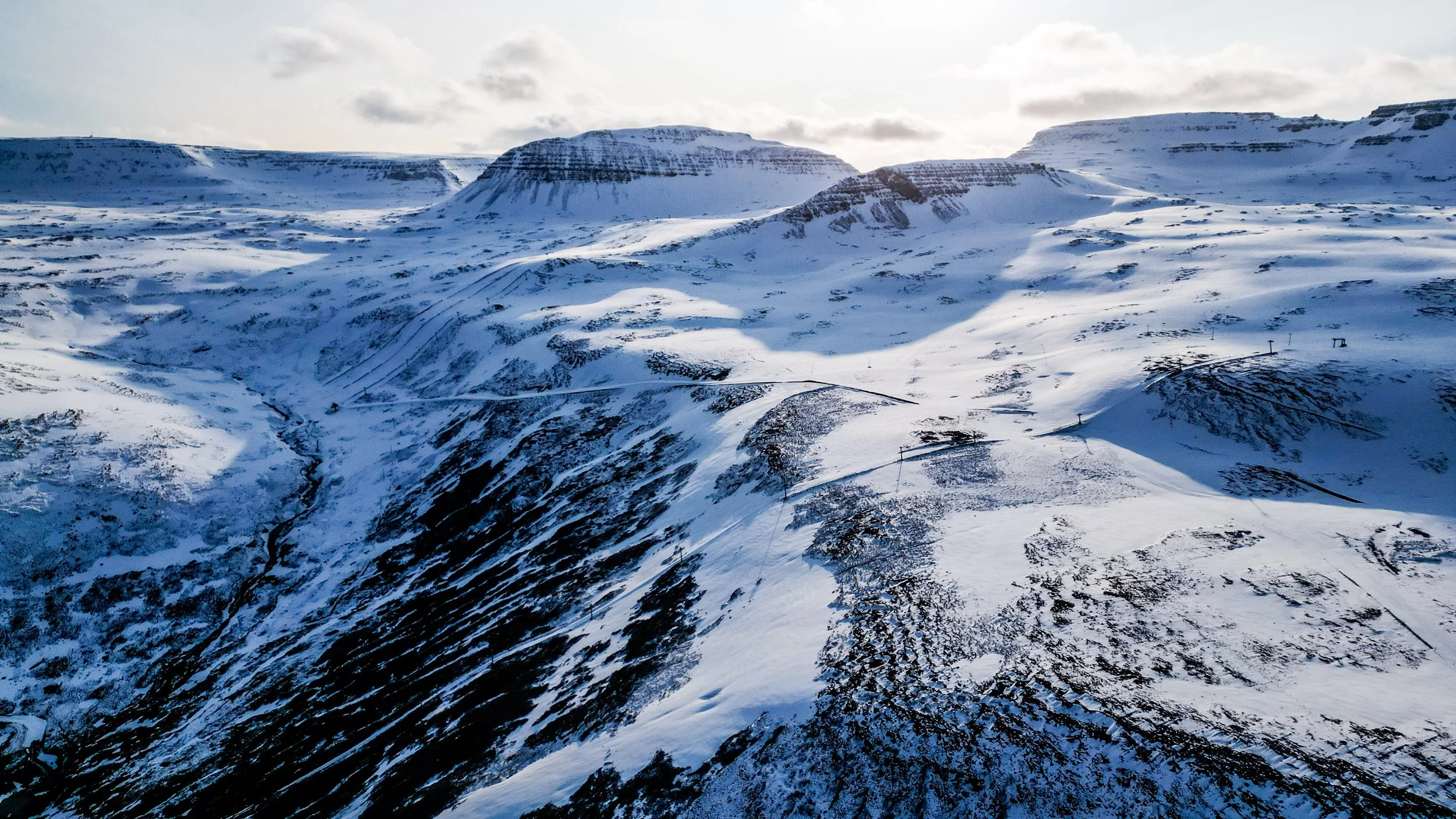 Snow-covered mountain landscape with ridges and slopes, cloudy sky overhead.