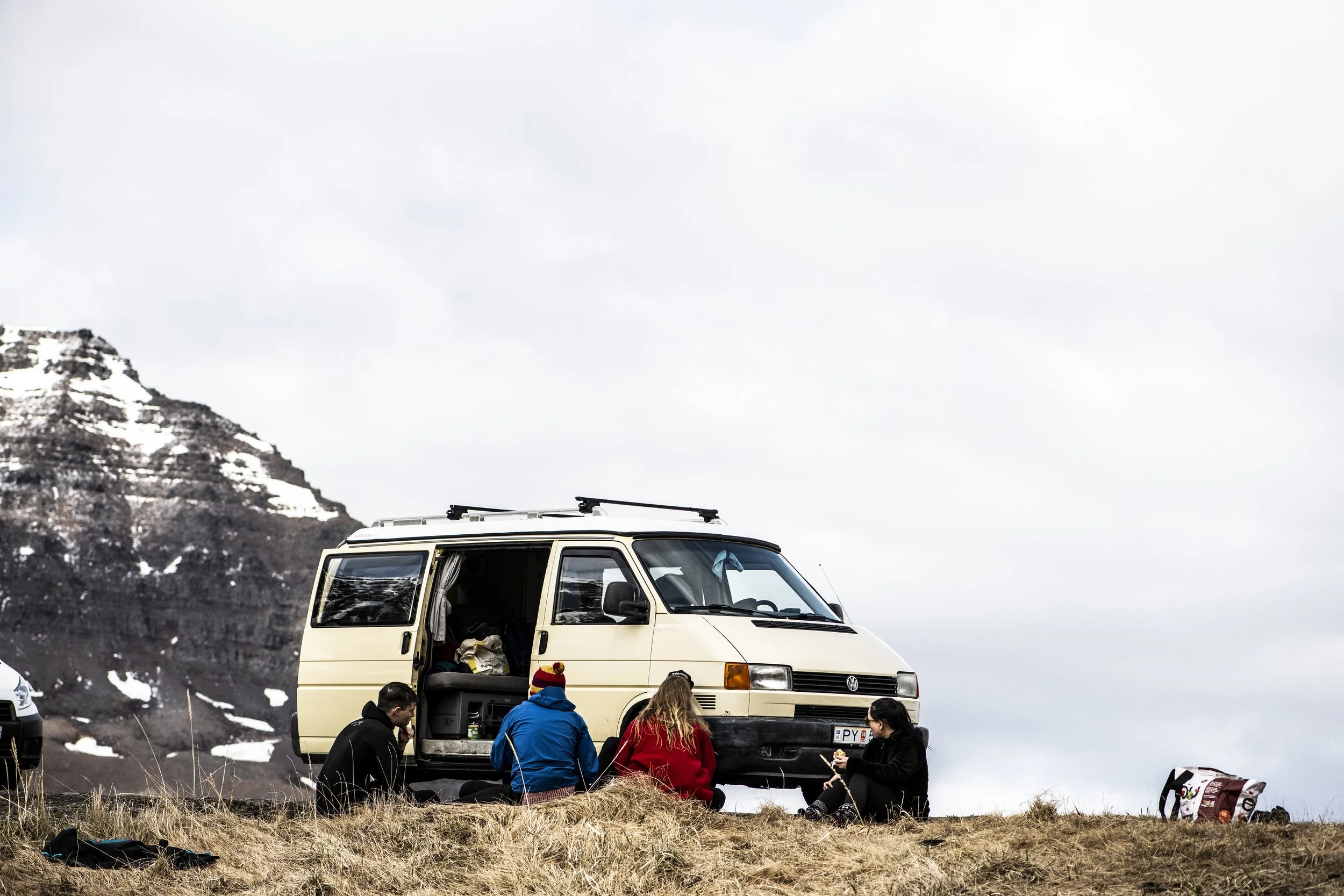 A group of people sitting and standing outside near a yellow van with a mountain and cloudy sky in the background during daytime.
