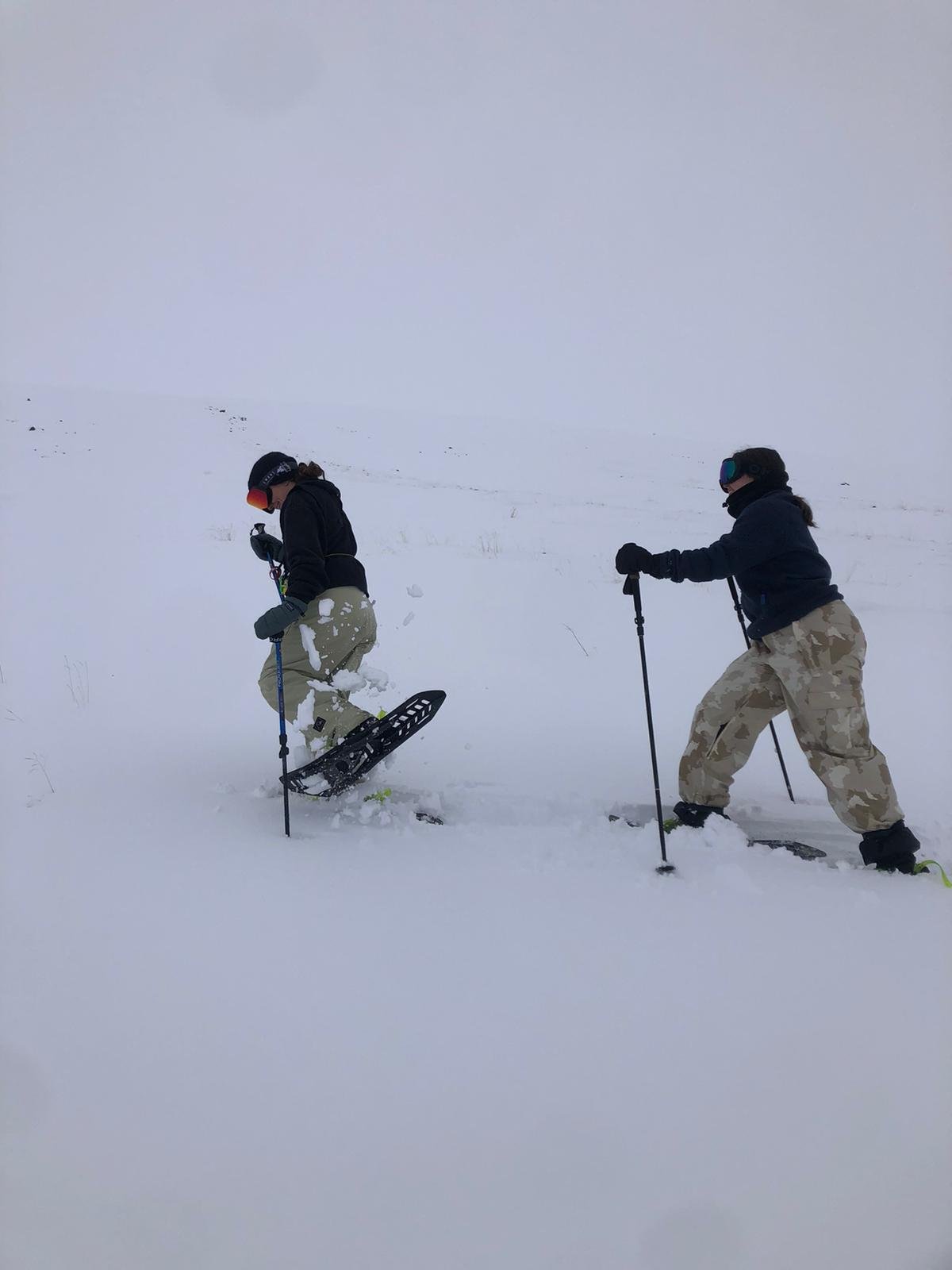 Two people snowboarding in a snowy landscape, dressed in winter gear with camouflage pants, using poles for support.