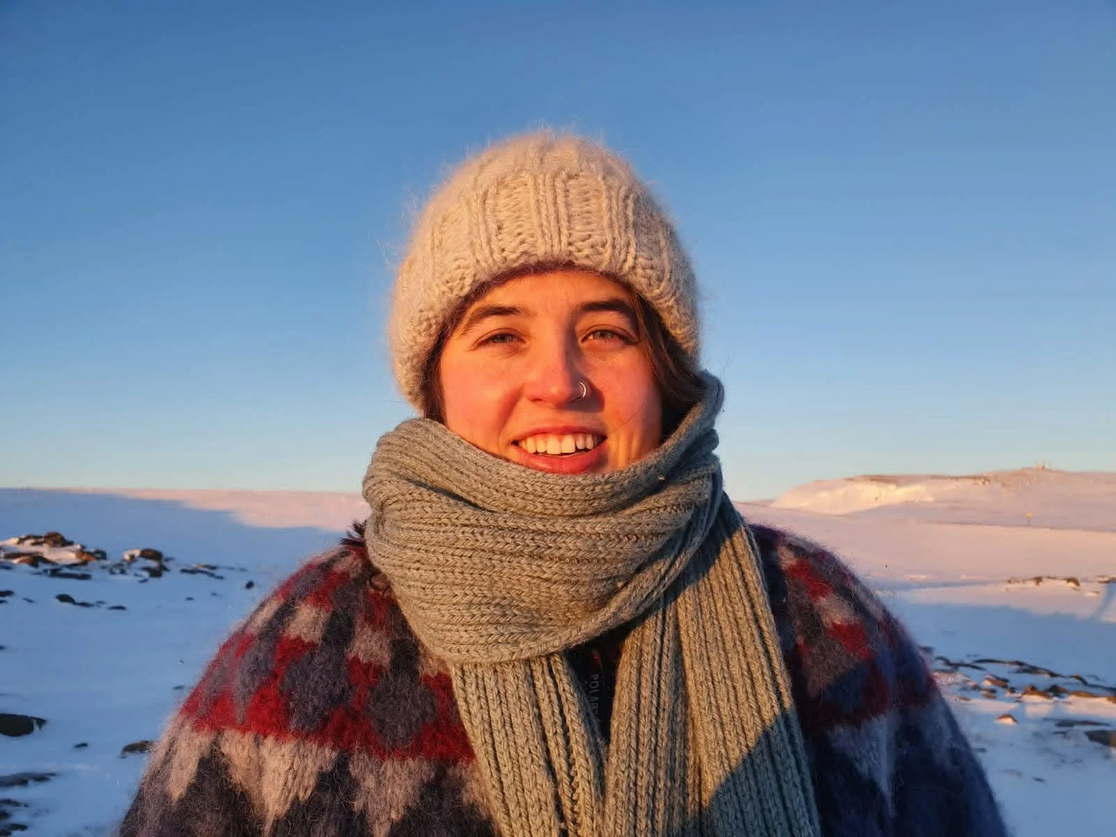 A smiling woman dressed warmly in a knit hat, scarf, and patterned sweater outdoors in a snowy landscape during sunset.