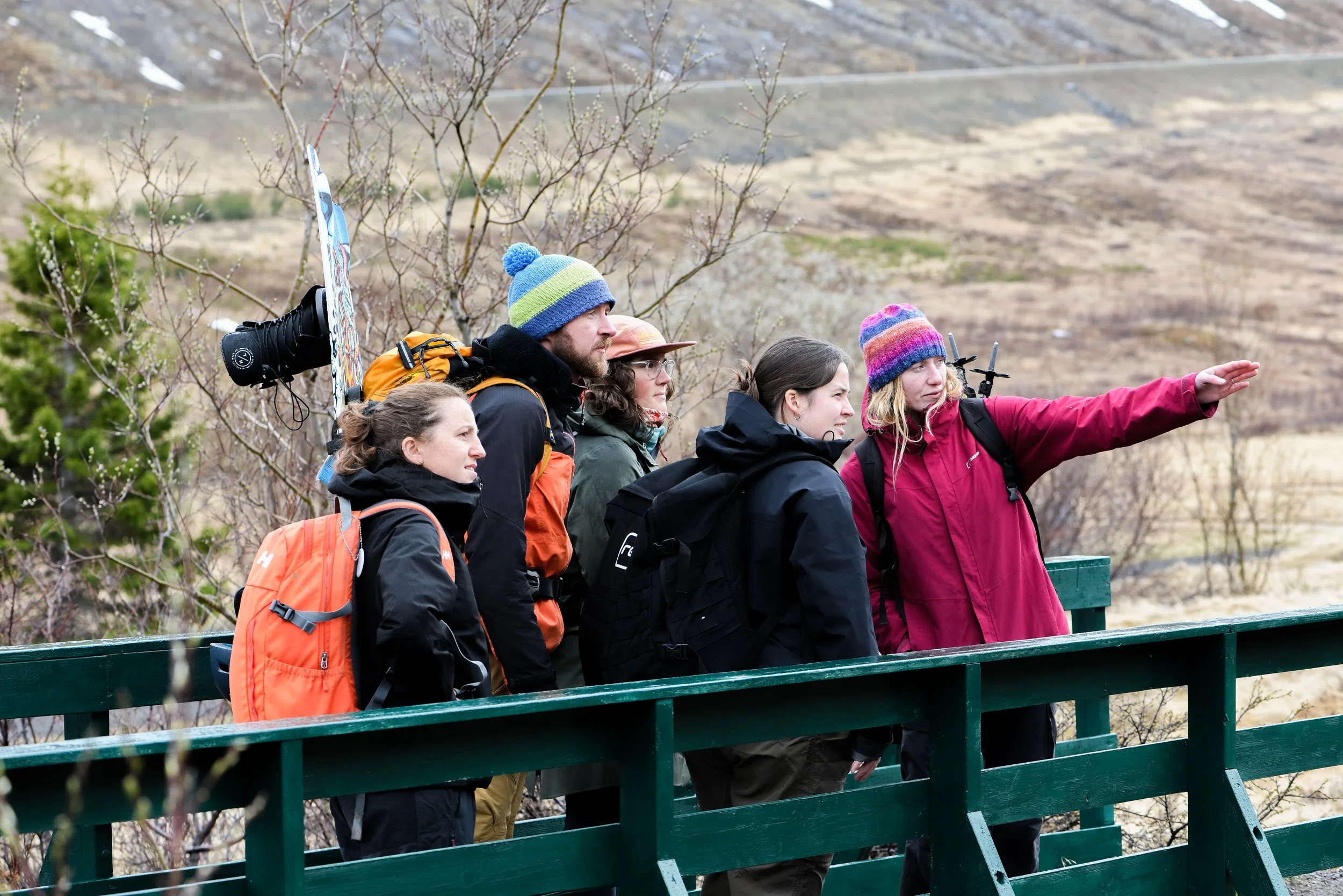 Group of five people in outdoor jackets and backpacks standing on a bridge, with one person pointing into the distance, set in a rural landscape with hills and sparse vegetation.