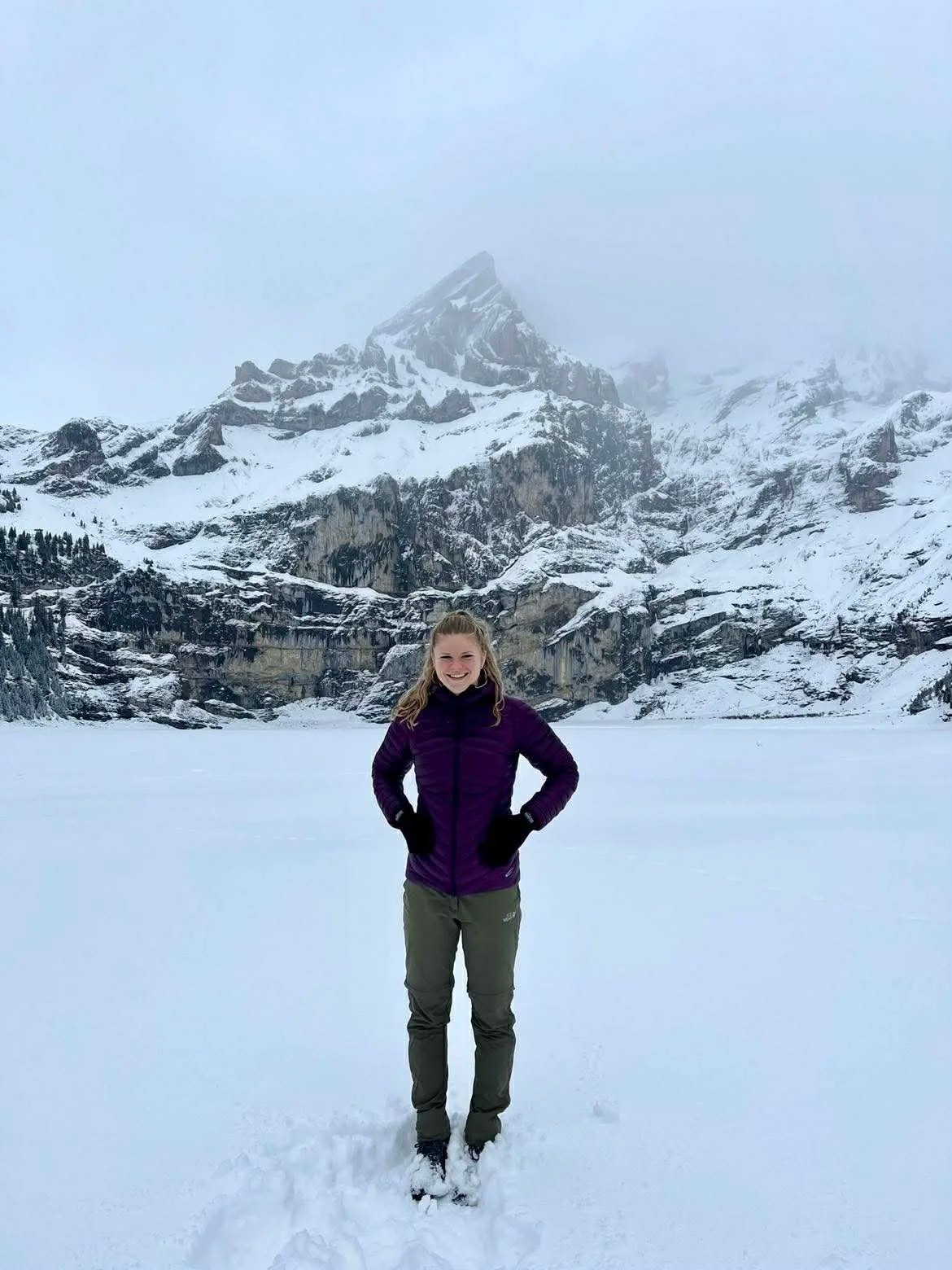 A young woman smiling in winter clothing standing on snow with mountains in the background, partially covered in snow and fog.