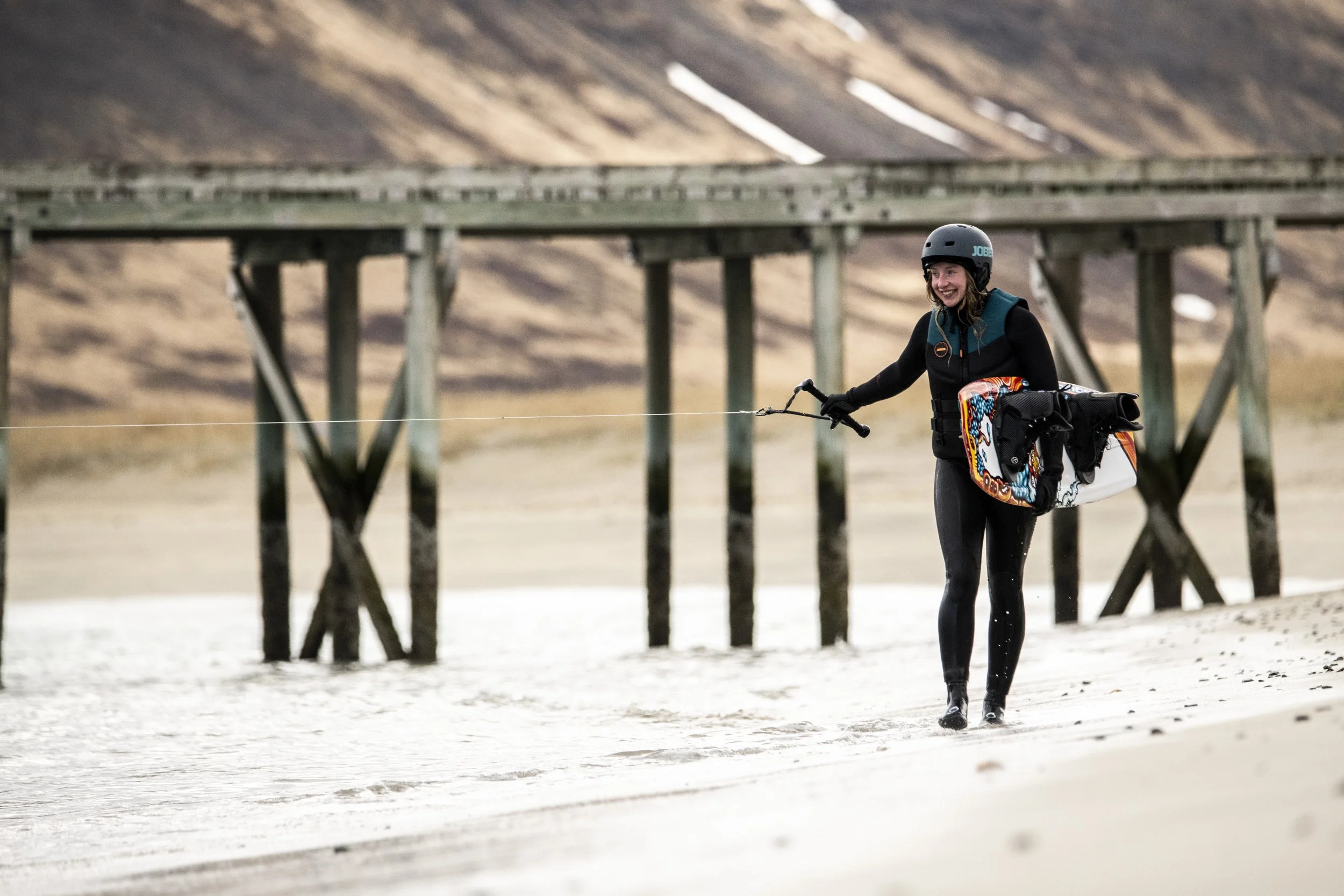 A woman in a wetsuit and helmet walking along a beach, carrying a kiteboarding kite, with a pier structure in the background and hills in the distance.