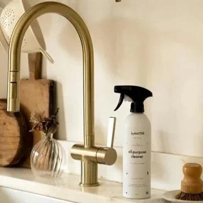 Kitchen sink with a brass faucet, a spray bottle of all-purpose cleaner, a wooden brush, and a small decorative vase with dried flowers.