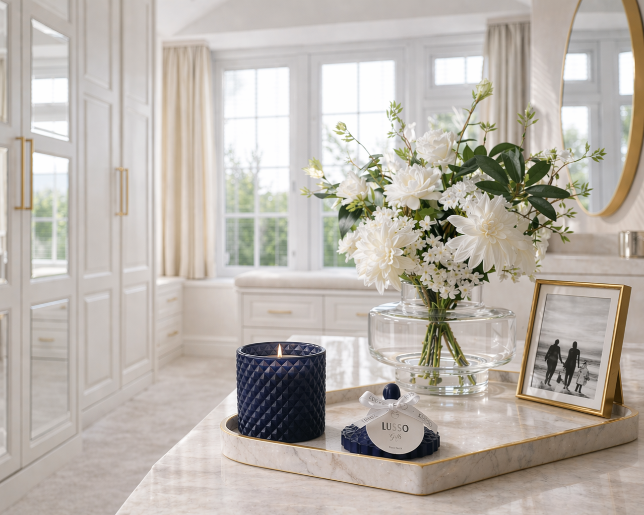 Decorative display on a marble countertop featuring a glass vase with white flowers, a black candle, a framed black-and-white family photo, and a small gift box with a white ribbon, in a bright room with large windows and elegant white cabinetry.