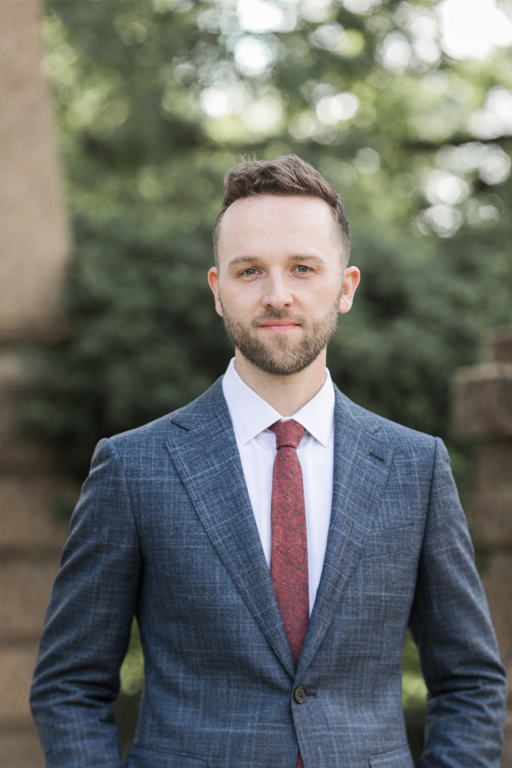 Geoff Wetrosky with short brown hair and a beard wearing a dark blue checkered suit, white shirt, and red tie standing outdoors with blurred green trees in the background.