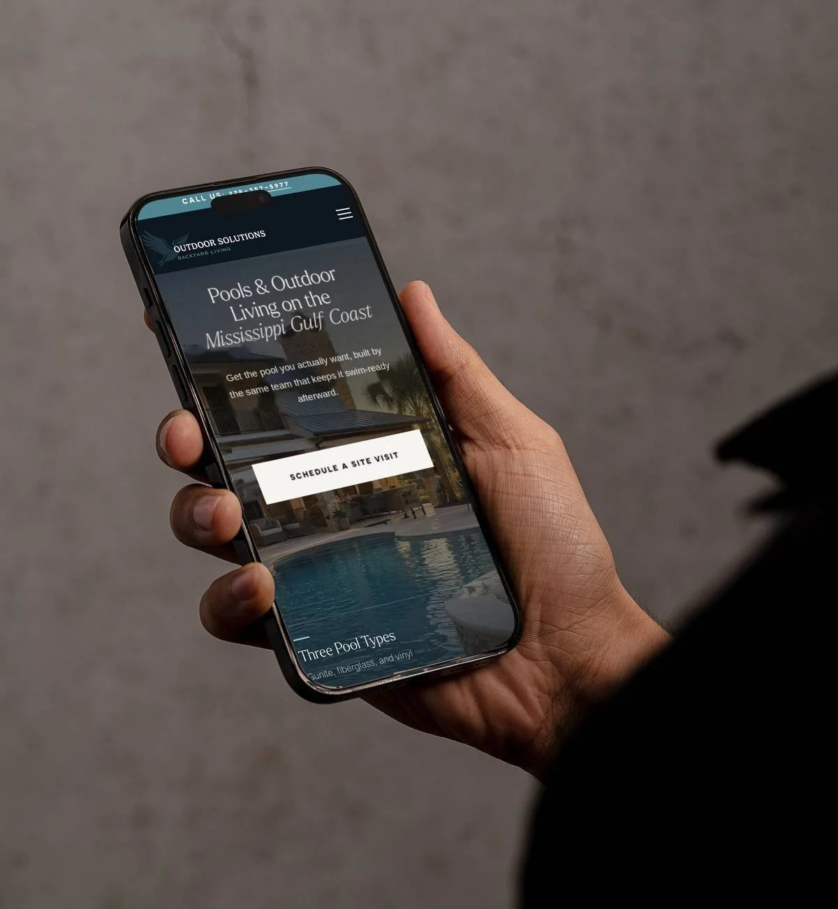 Hand holding a smartphone displaying an outdoor pool design website for Mississippi Gulf Coast living.