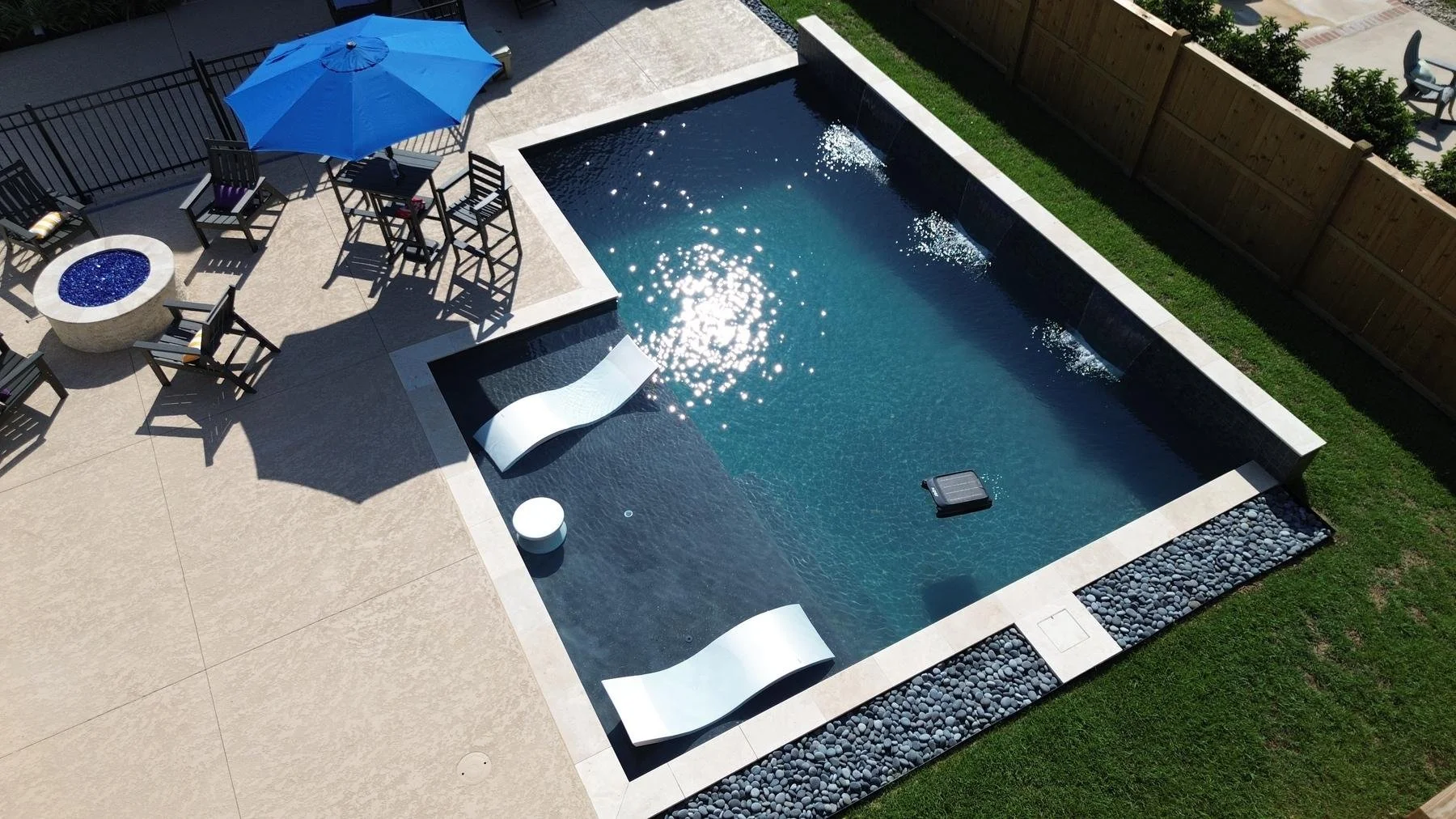 Aerial view of a modern backyard pool with lounge chairs, blue umbrella, fire pit, and landscaped stone border
