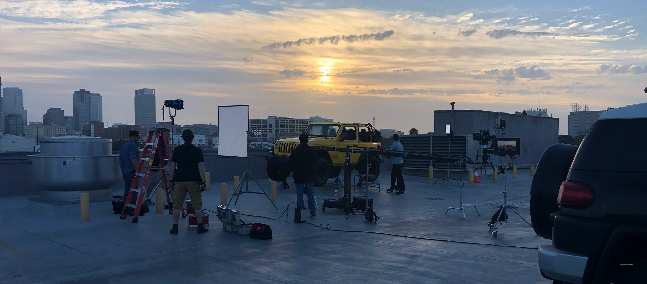 A behind-the-scenes view of a film or photo shoot on a rooftop at sunset, featuring crew members, a yellow off-road vehicle, professional equipment, and city skyline in the background.