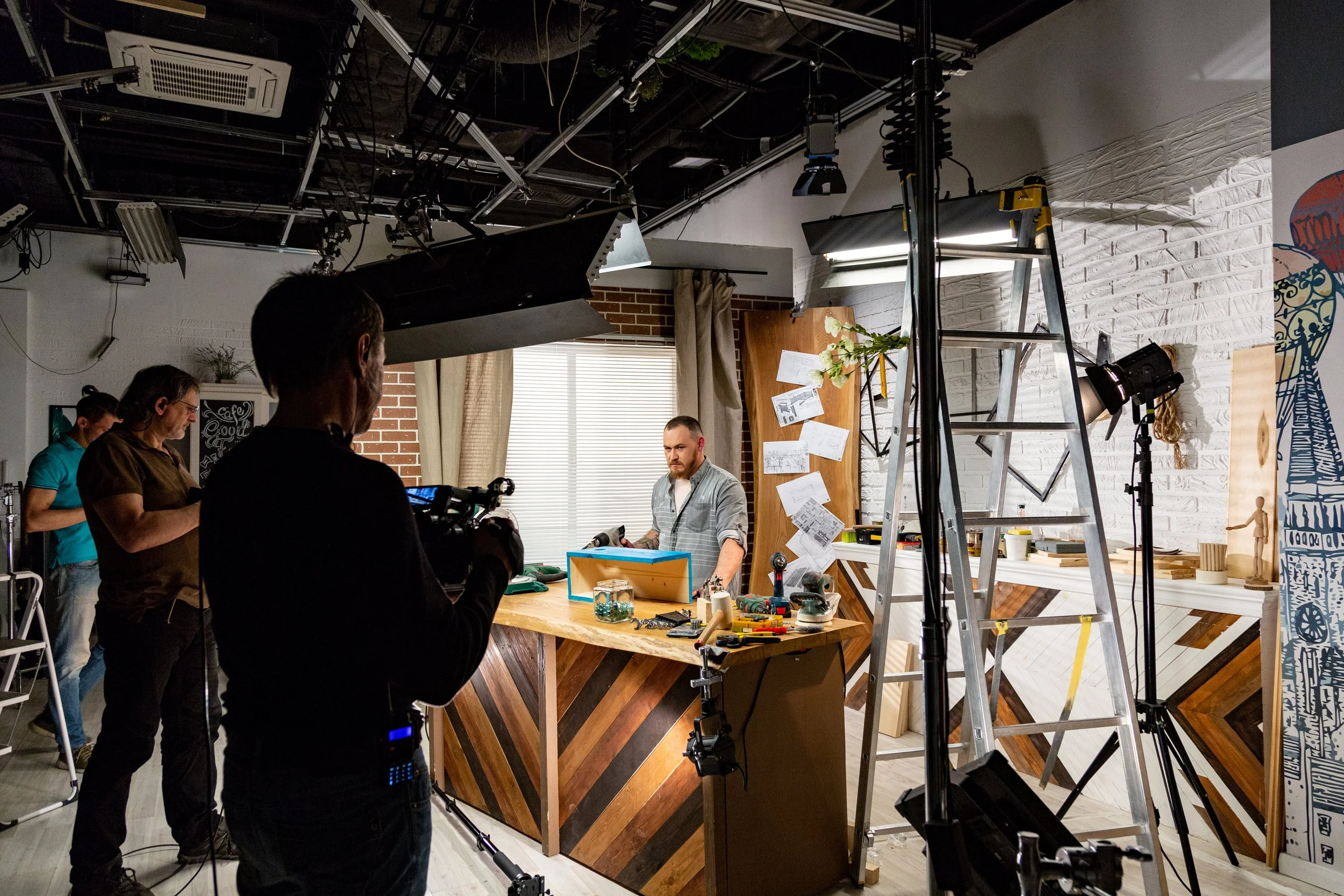 Set of a video shoot or filming studio with crew members, camera equipment, ladder, and a man standing behind a wooden counter in front of a white brick wall with various papers and decorations.