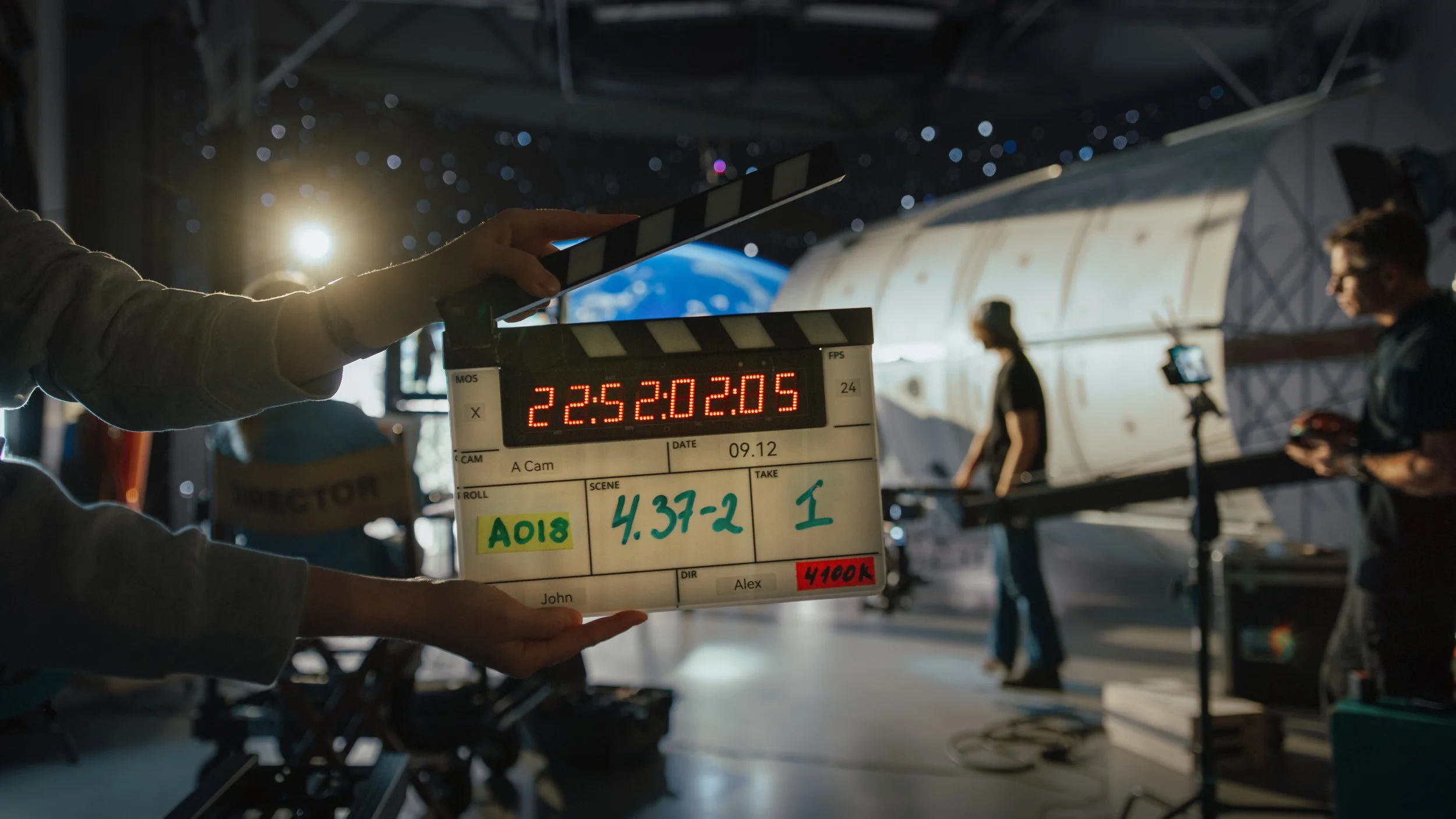 Inside a space station or spacecraft, crew members prepare for a scene or shot with a clapperboard, with Earth visible through a window and a galaxy or stars outside.