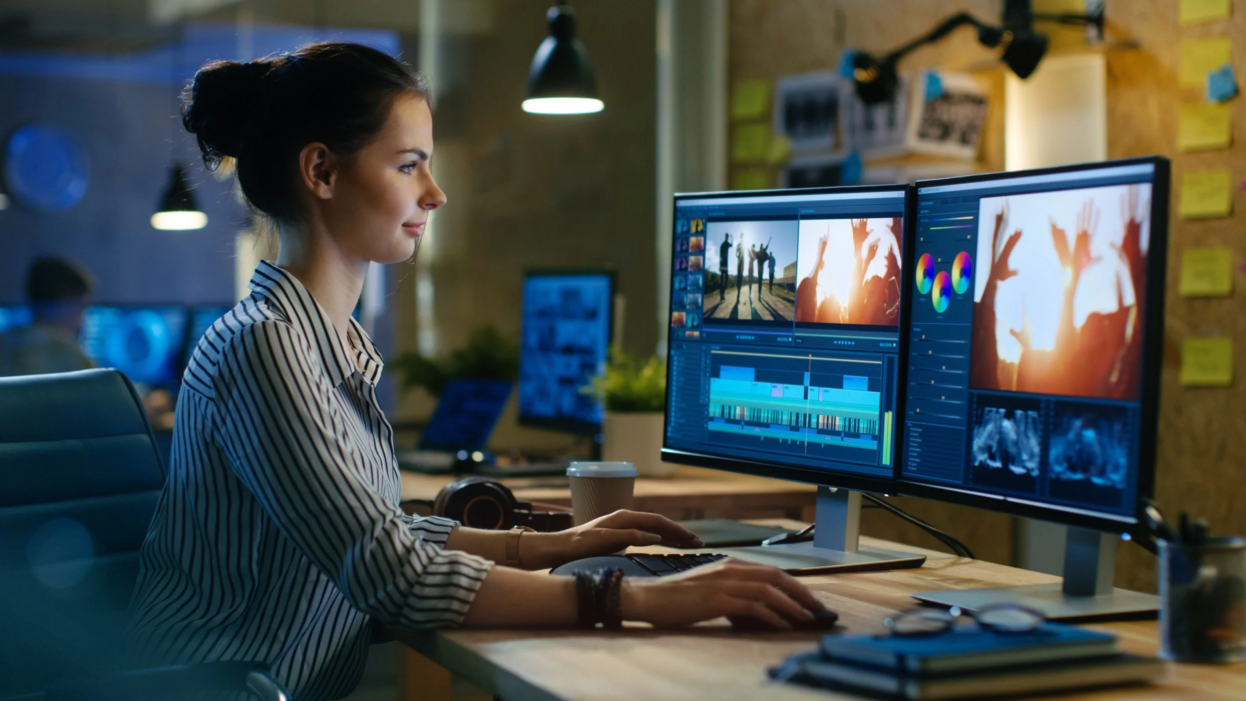 A woman working on video editing at a desk with two computer monitors showing video editing software.