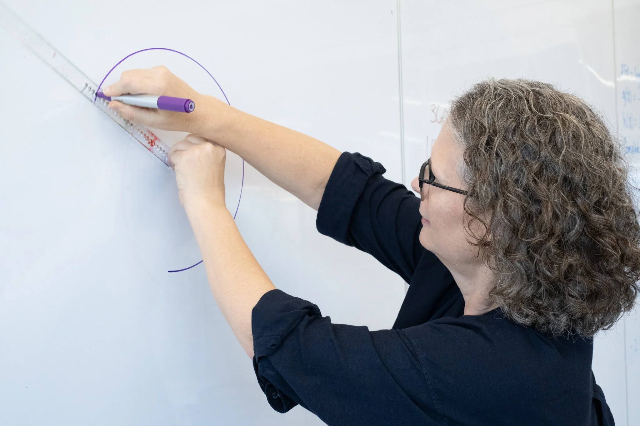 A woman with glasses and curly hair is drawing on a whiteboard using a purple marker and a ruler-like tool called a goround.
