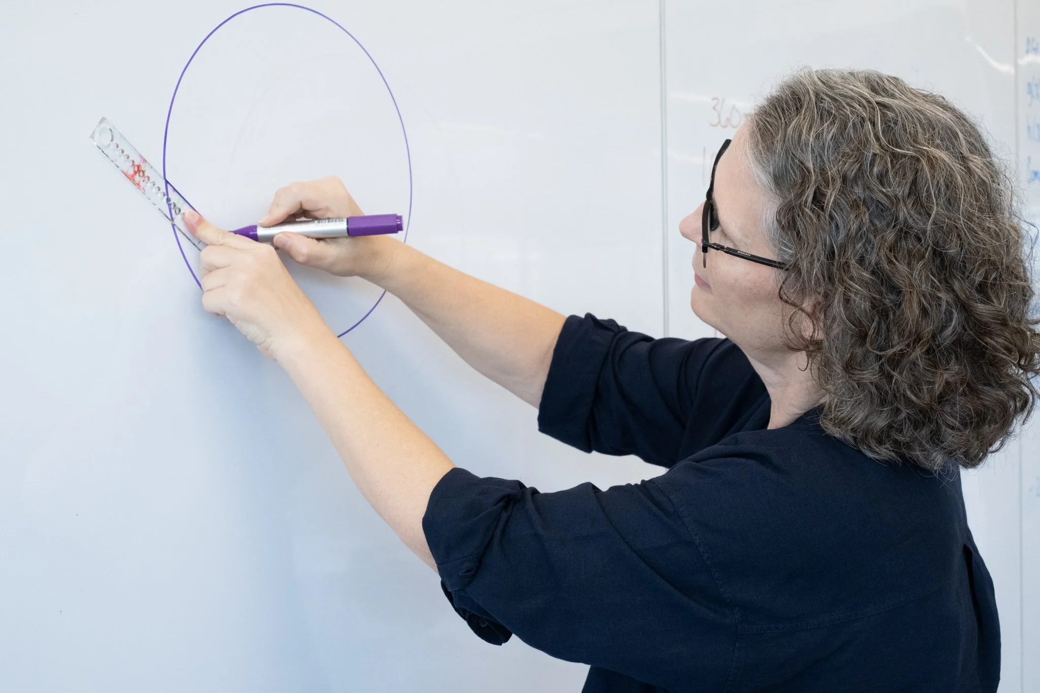 A woman with curly hair and glasses drawing a chord across a circle with purple marker on a whiteboard, using a ruler-like tool called a goround.