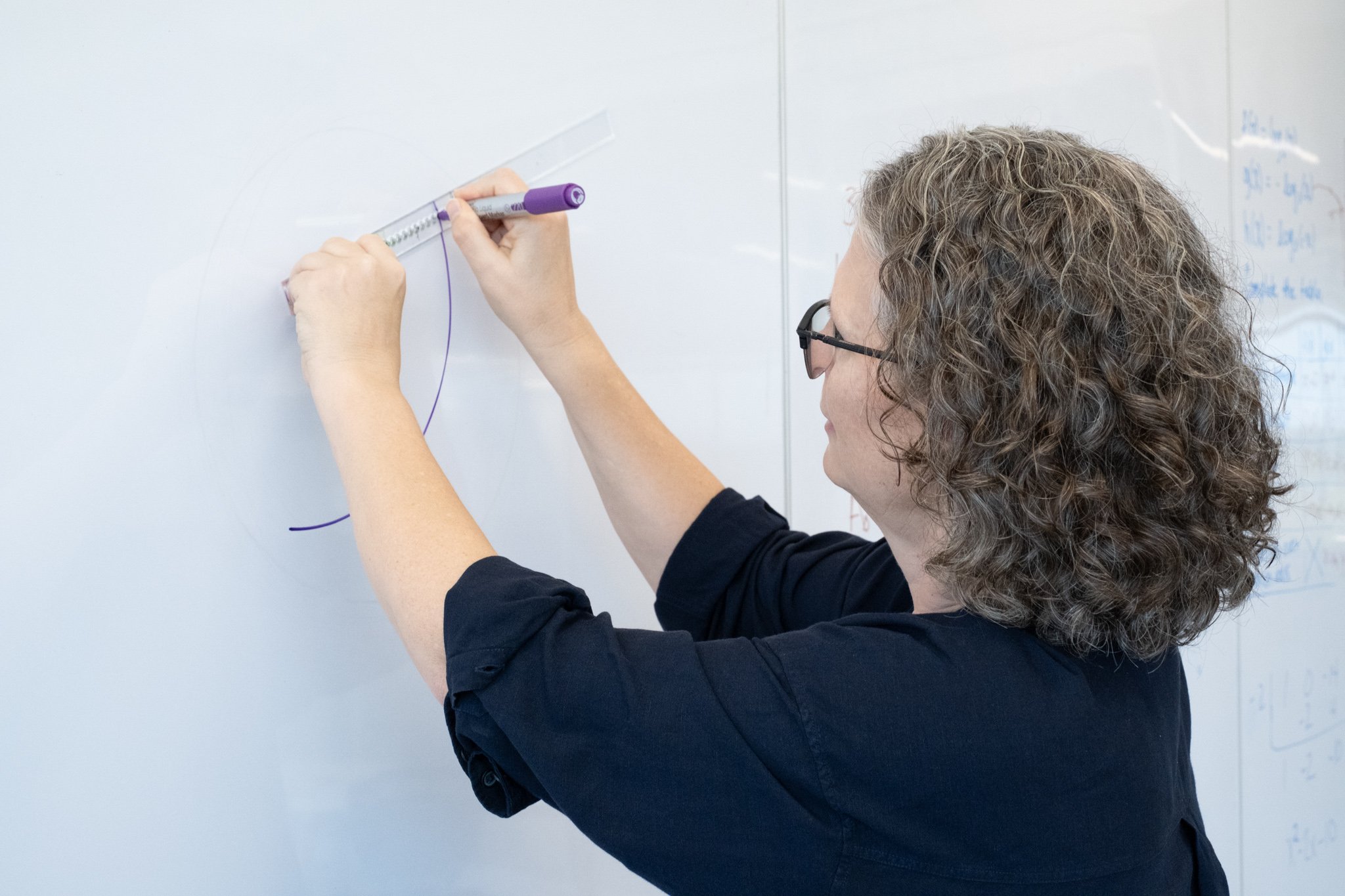 A woman with curly hair and glasses making a circle on a whiteboard with a purple marker.