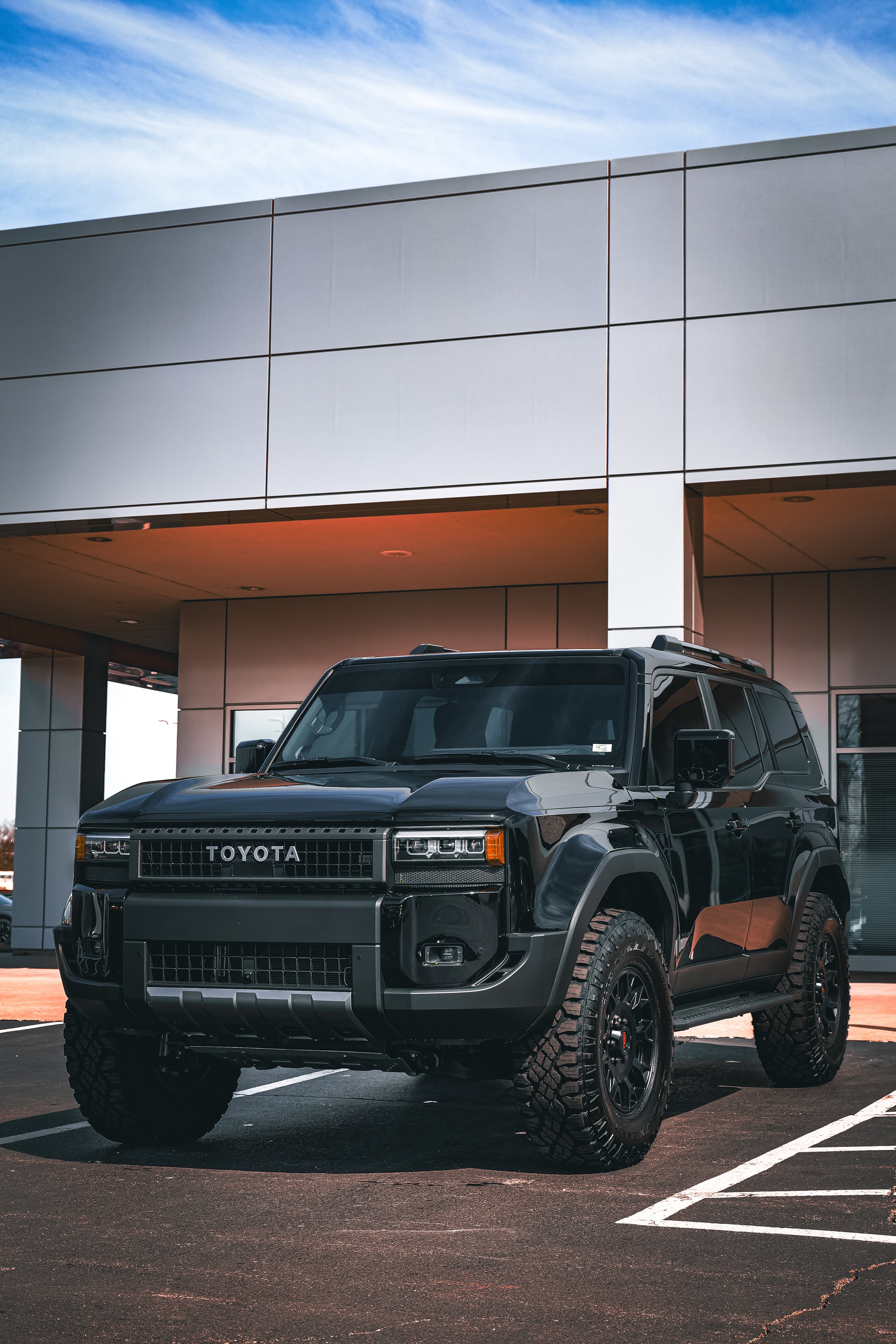 A black Toyota SUV parked in front of a modern building with large gray panels and a partly cloudy sky.