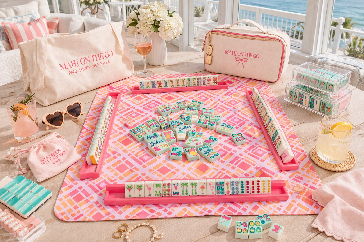 A colorful mahjong game setup on a pink and orange patterned tablecloth with decorative items, glasses of drinks, and storage bags, in a bright room with a view of the ocean outside.