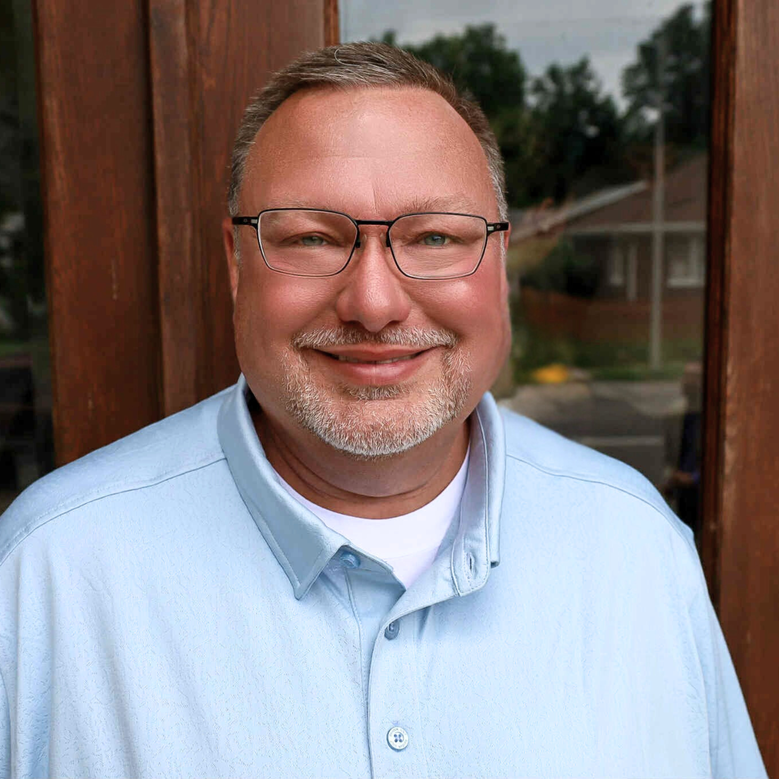 Close-up of a smiling middle-aged man wearing glasses, a light blue collared shirt, standing outdoors near a wooden fence with trees and a cloudy sky in the background.