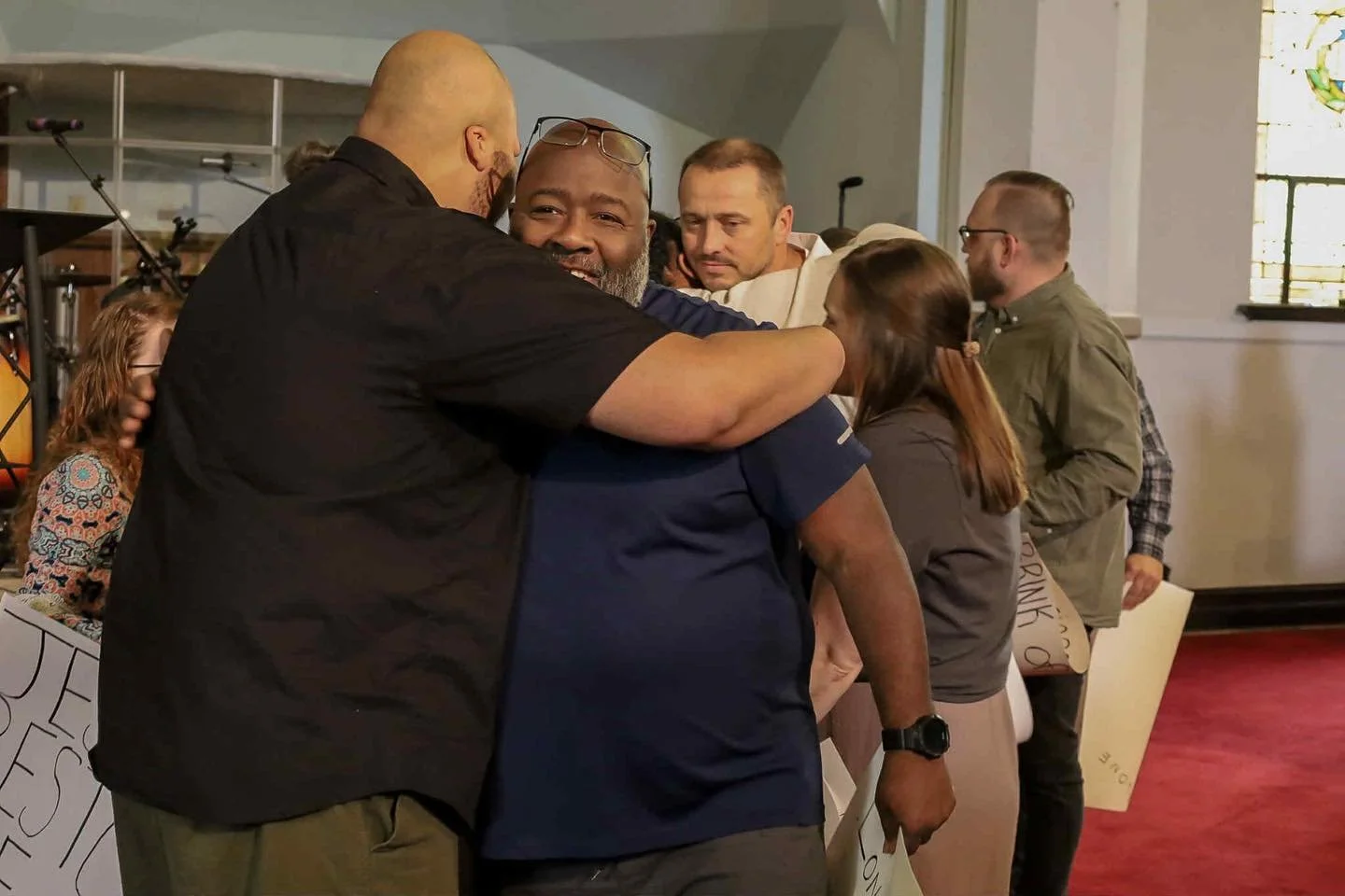 A group of people sharing a hug and smiling in a church or community hall. Some are holding signs with partially visible text.