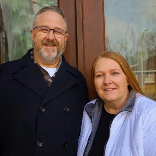 A man with glasses, grey hair, and a beard wearing a navy coat, standing next to a woman with red hair and fair skin wearing a light jacket. They are standing outside in front of a glass window.