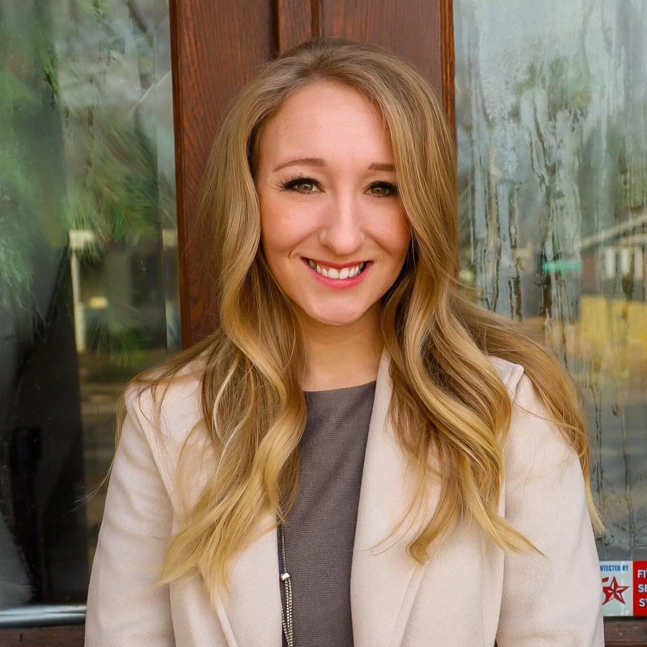 A young woman with long, wavy blonde hair, smiling and wearing a beige coat and gray top, standing outside in front of a glass window with reflections.