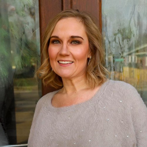 A woman with shoulder-length blonde hair smiling outdoors near a window.