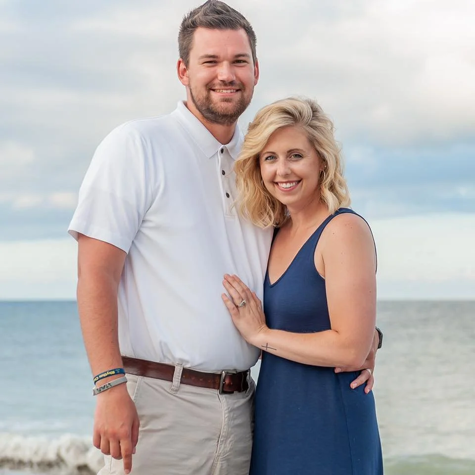 A smiling couple stands close together on a beach with ocean in the background.