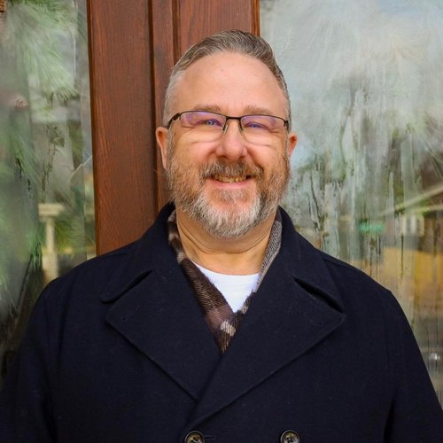 A smiling man with glasses, a beard, and short hair posing outside near a wooden and glass structure.