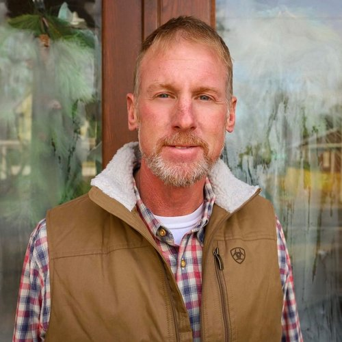 A middle-aged man with blond hair and a beard standing outside in front of a glass window with greenery inside. He is wearing a plaid shirt and a brown vest.