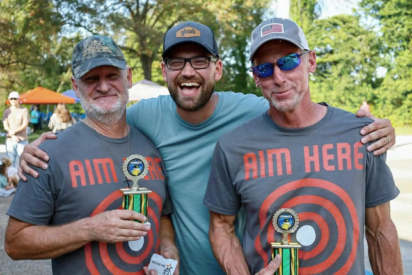 Three men smiling at an outdoor event, two wearing gray t-shirts with the words 'AIM HERE' and target symbols, holding trophies, with trees and tents in the background.